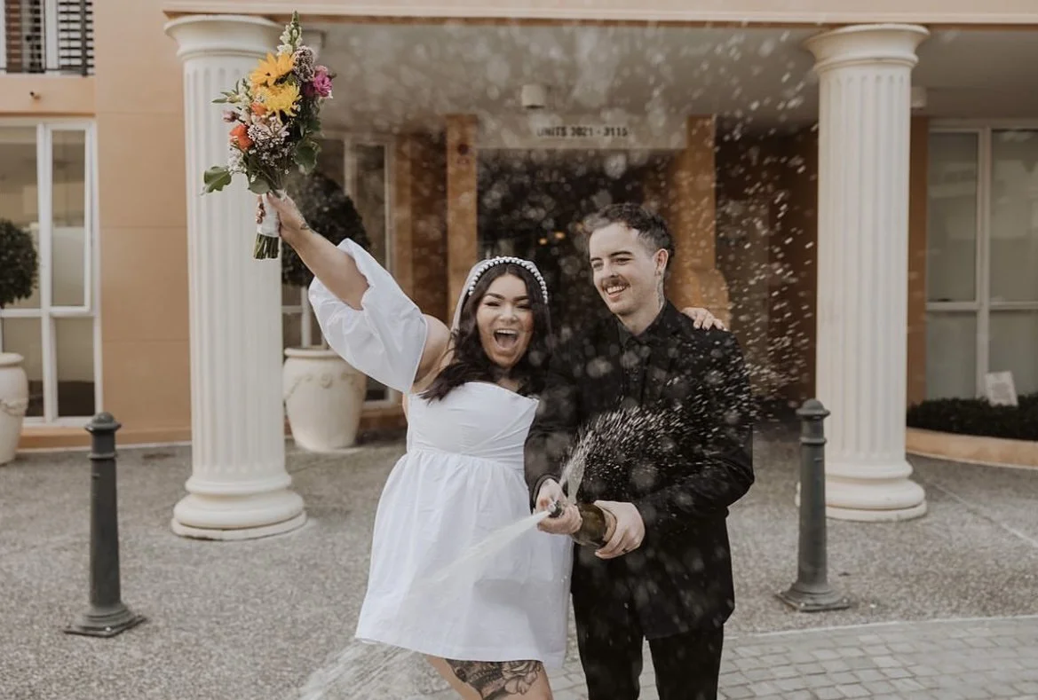 A joyful couple celebrates outside a building, with the woman wearing a white dress and holding flowers, and the man dressed in black popping a champagne bottle.