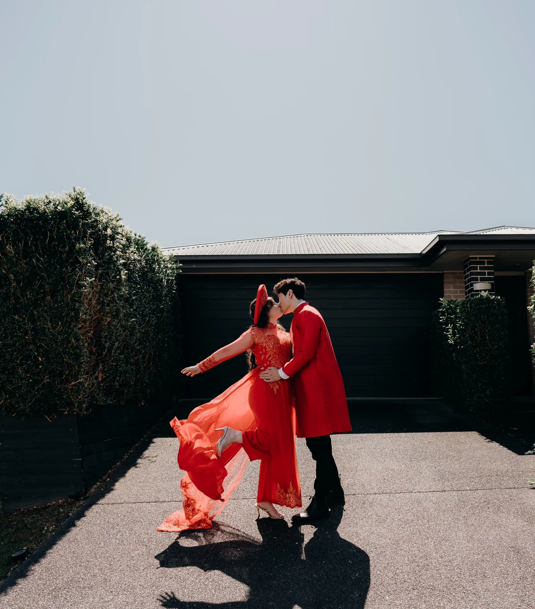 A couple in red attire kissing in front of a house, with one person wearing a traditional dress and hat, and the other in a long coat.