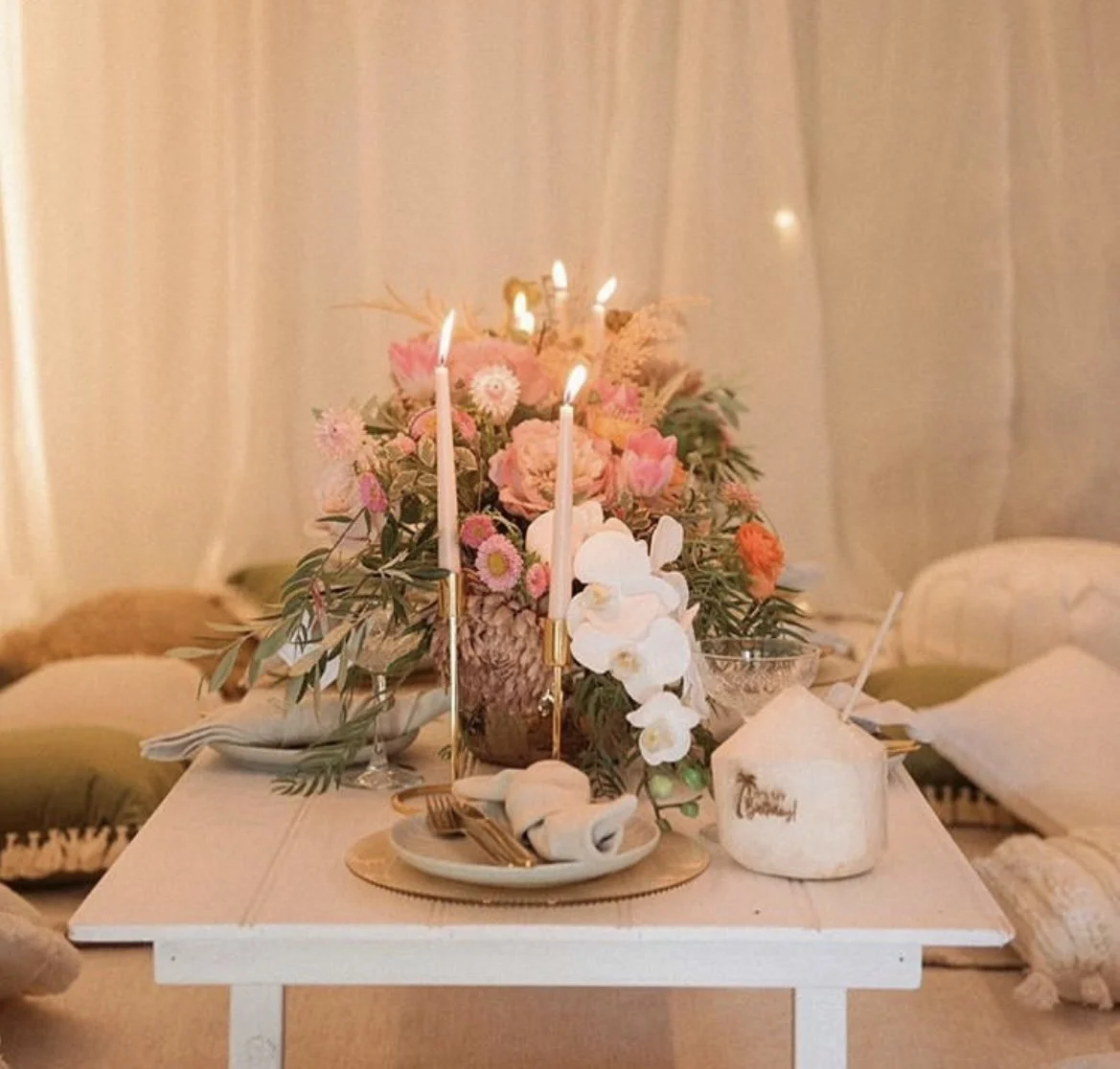 Elegant table setting with floral centerpiece, lit candles, and plates with cutlery on a white table. A coconut drink is placed beside the setting, against a soft backdrop.