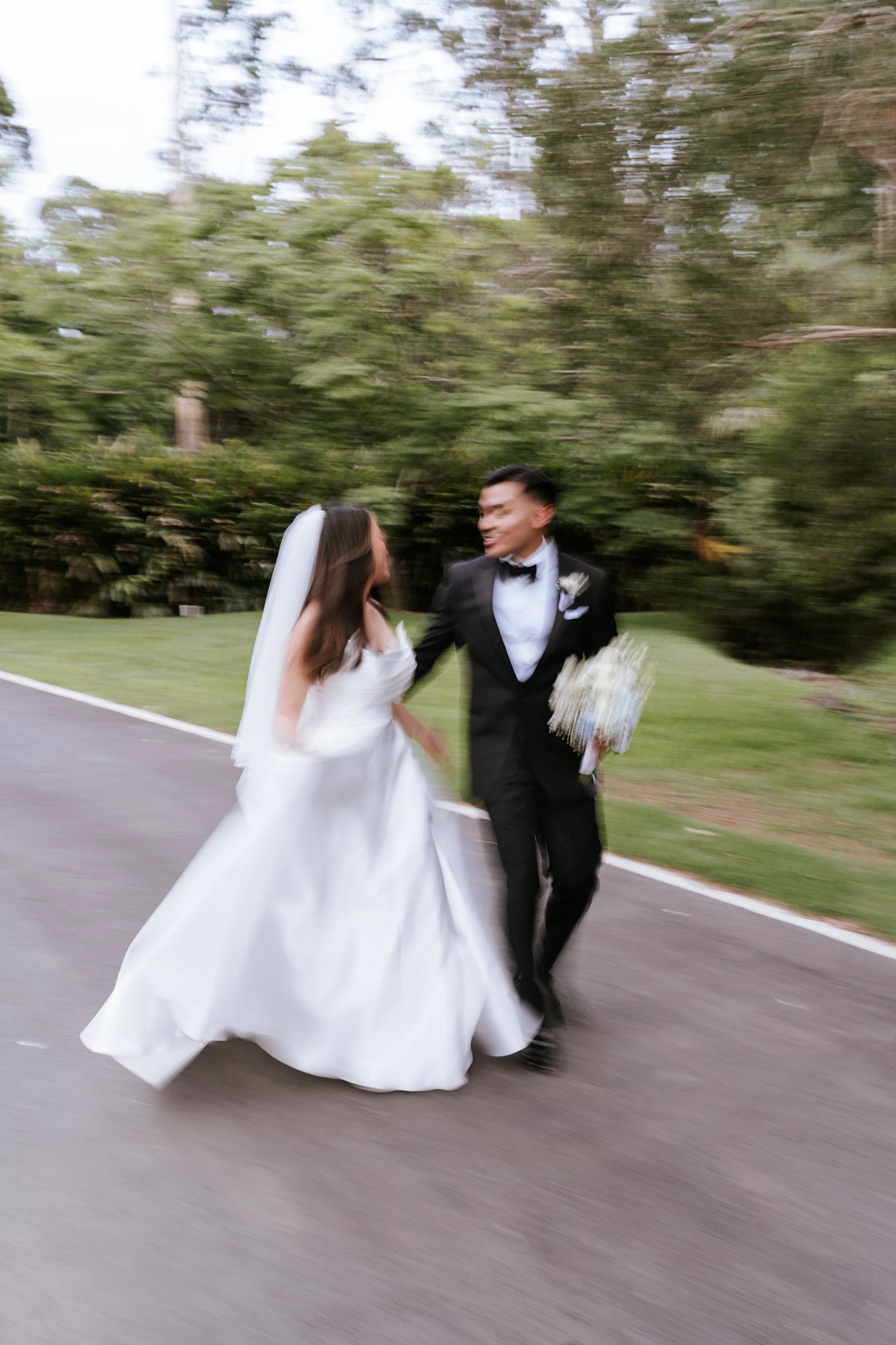 Bride and groom in formal attire walking on a path with blurred motion, surrounded by greenery.