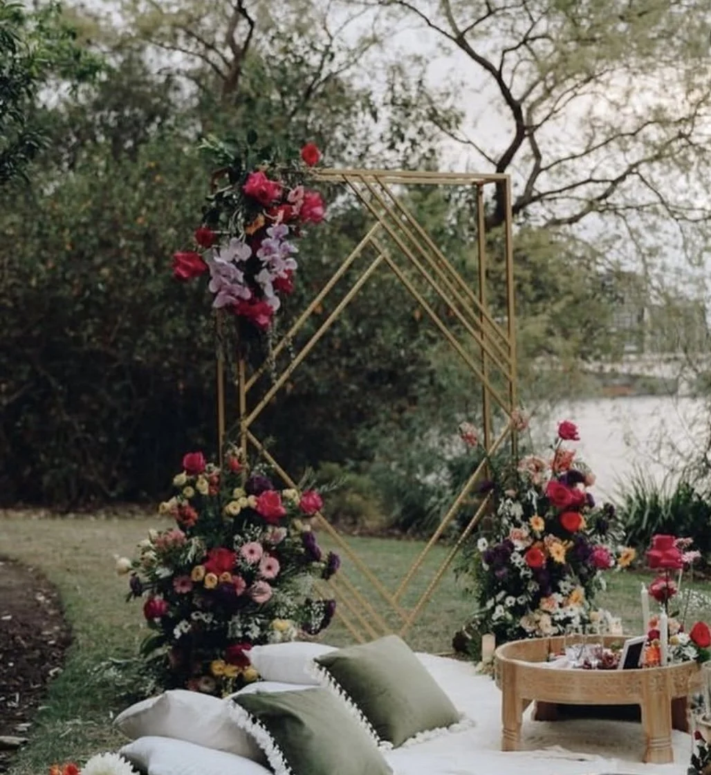 Outdoor bohemian wedding setup with gold geometric arch, floral arrangements, cushions, and a wooden table on grass.