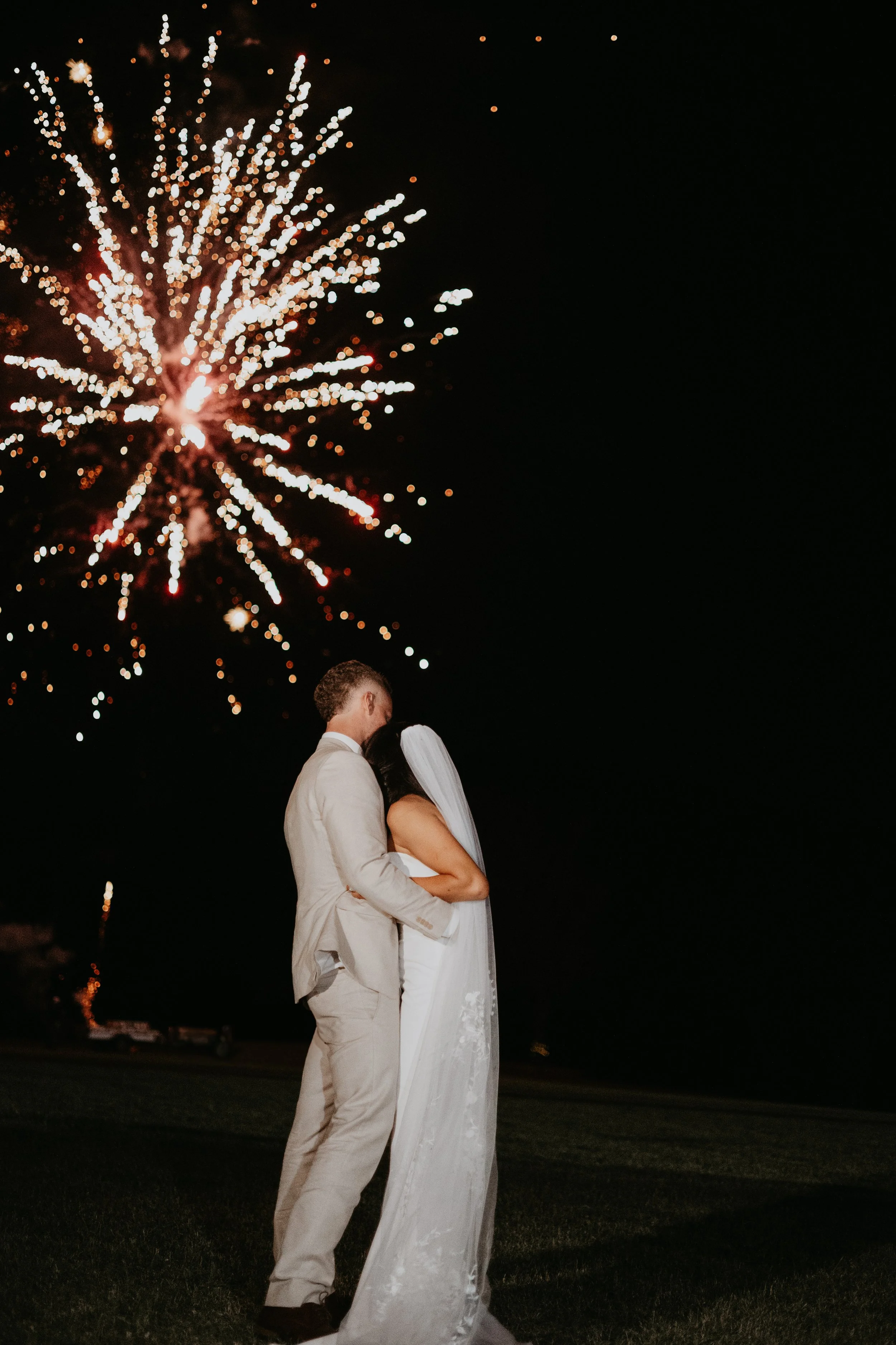 Bride and groom embracing under fireworks at night.