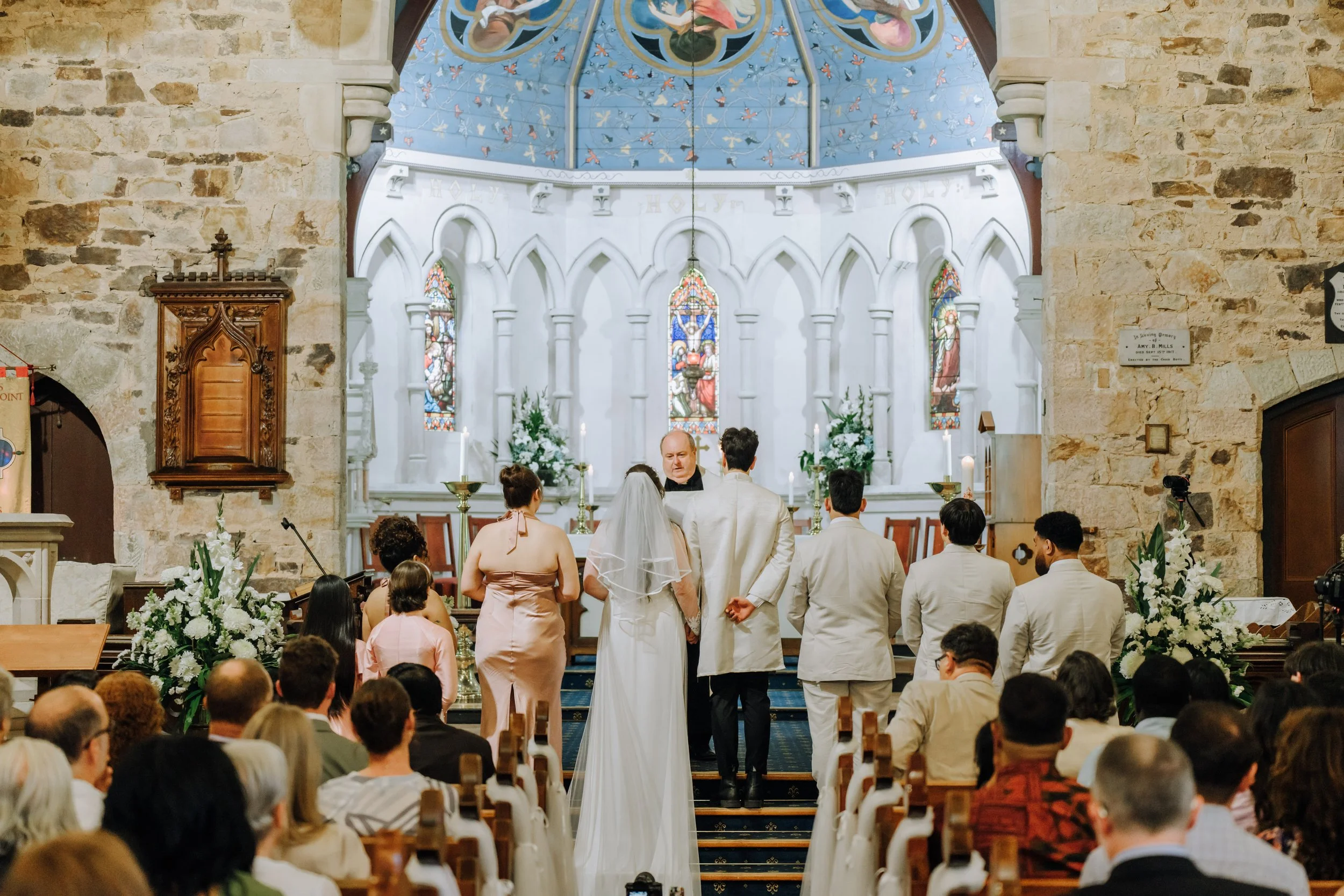 A wedding ceremony in a church with a bride and groom standing at the altar, surrounded by bridesmaids and groomsmen. Guests are seated, watching the ceremony. The church interior features stone walls and stained glass windows.