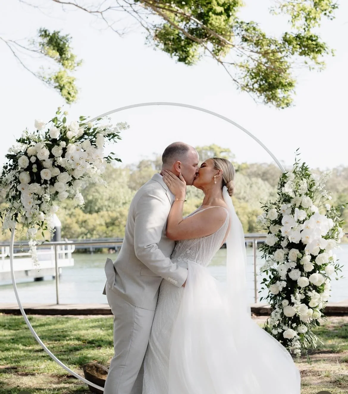 Bride and groom kissing outdoors under a floral arch during their wedding ceremony.