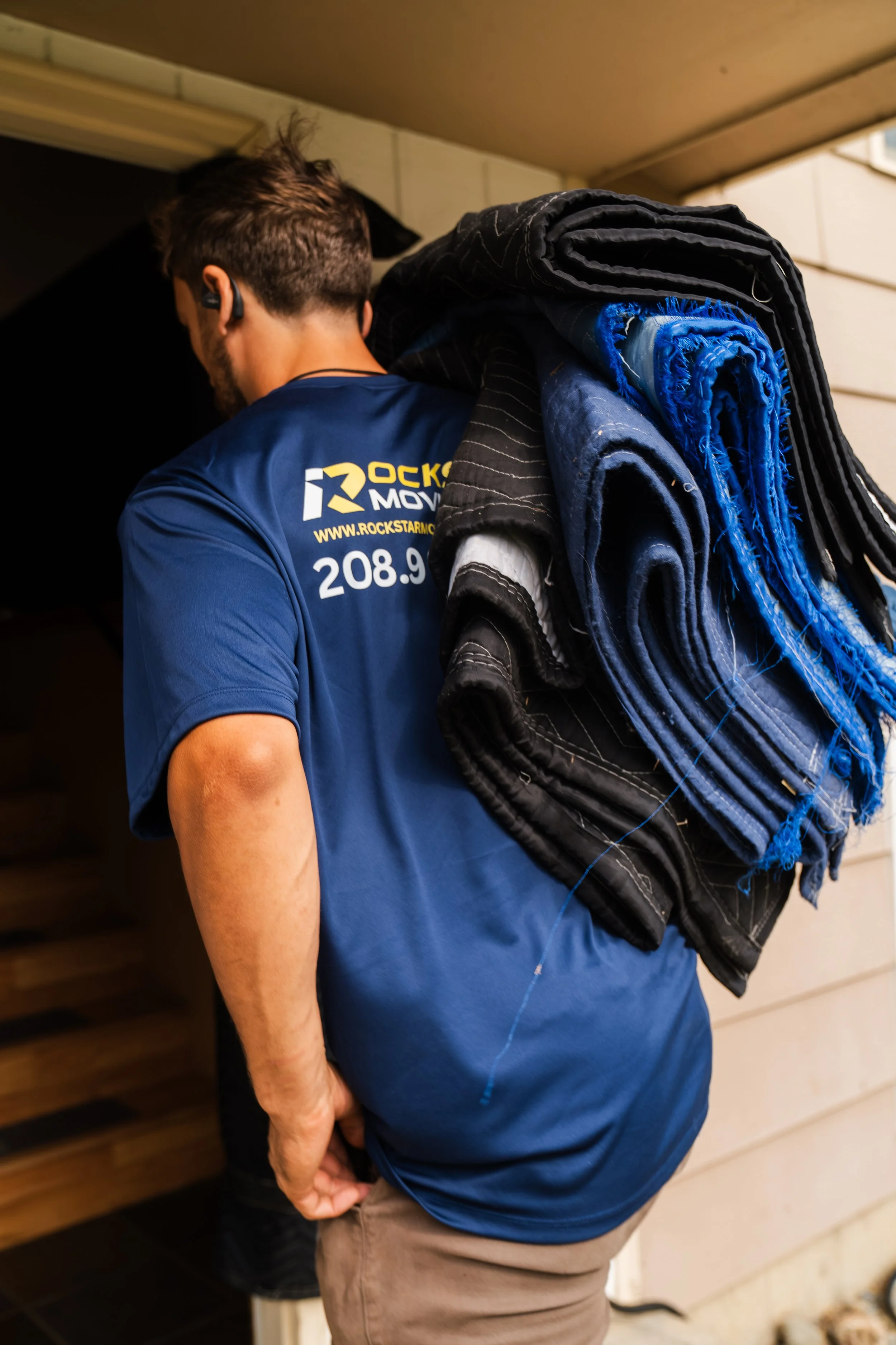 A man carrying rolled-up fabrics or blankets on his shoulder, wearing a blue Rockstar Moving Company t-shirt, standing near a wooden entrance door.