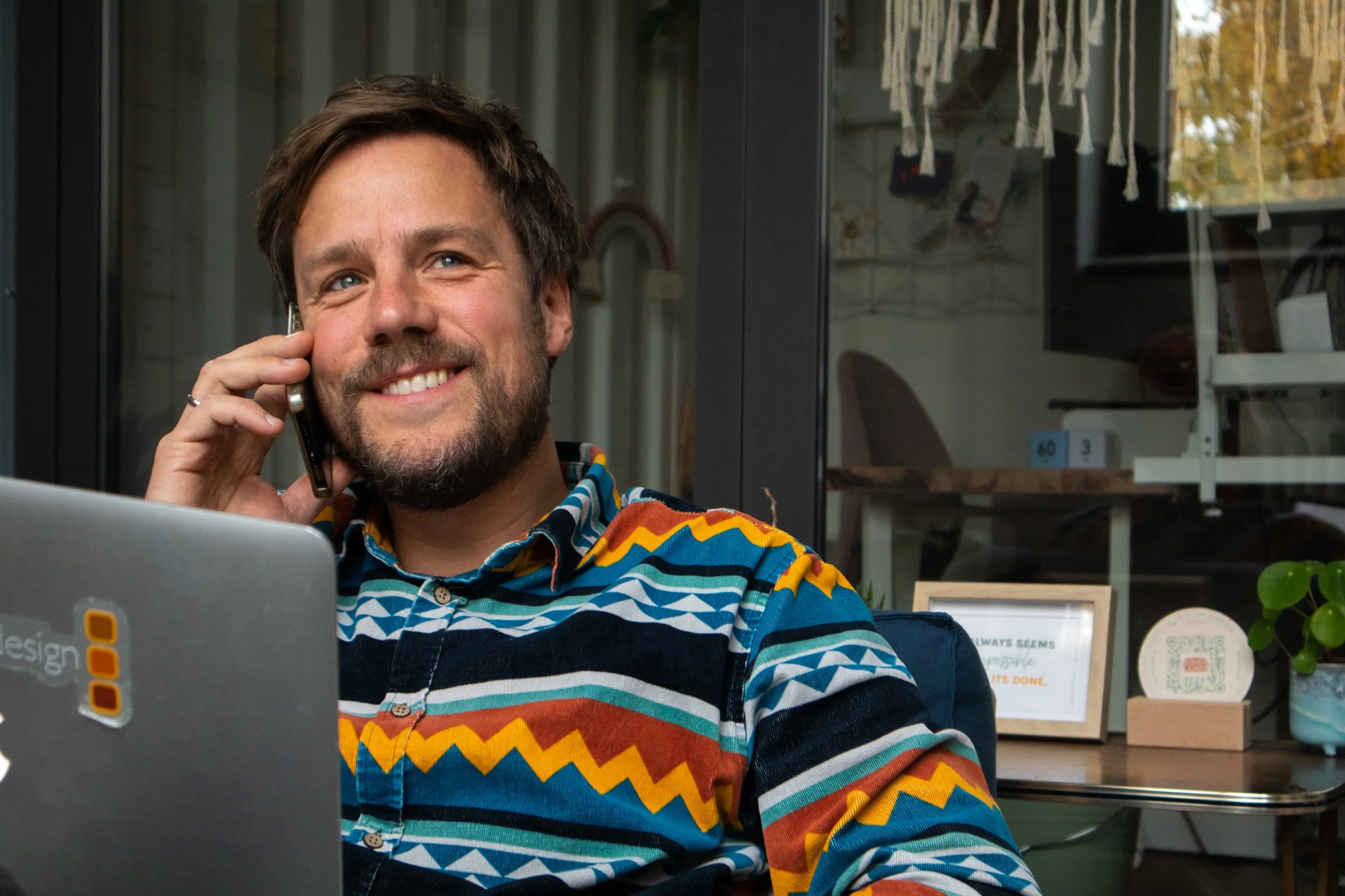Rob sitzt draußen, spricht am Handy und hat einen Laptop vor sich, trägt ein bunt gestreiftes Hemd, im Hintergrund sind Regale, Pflanzen und eine Tafel sichtbar.