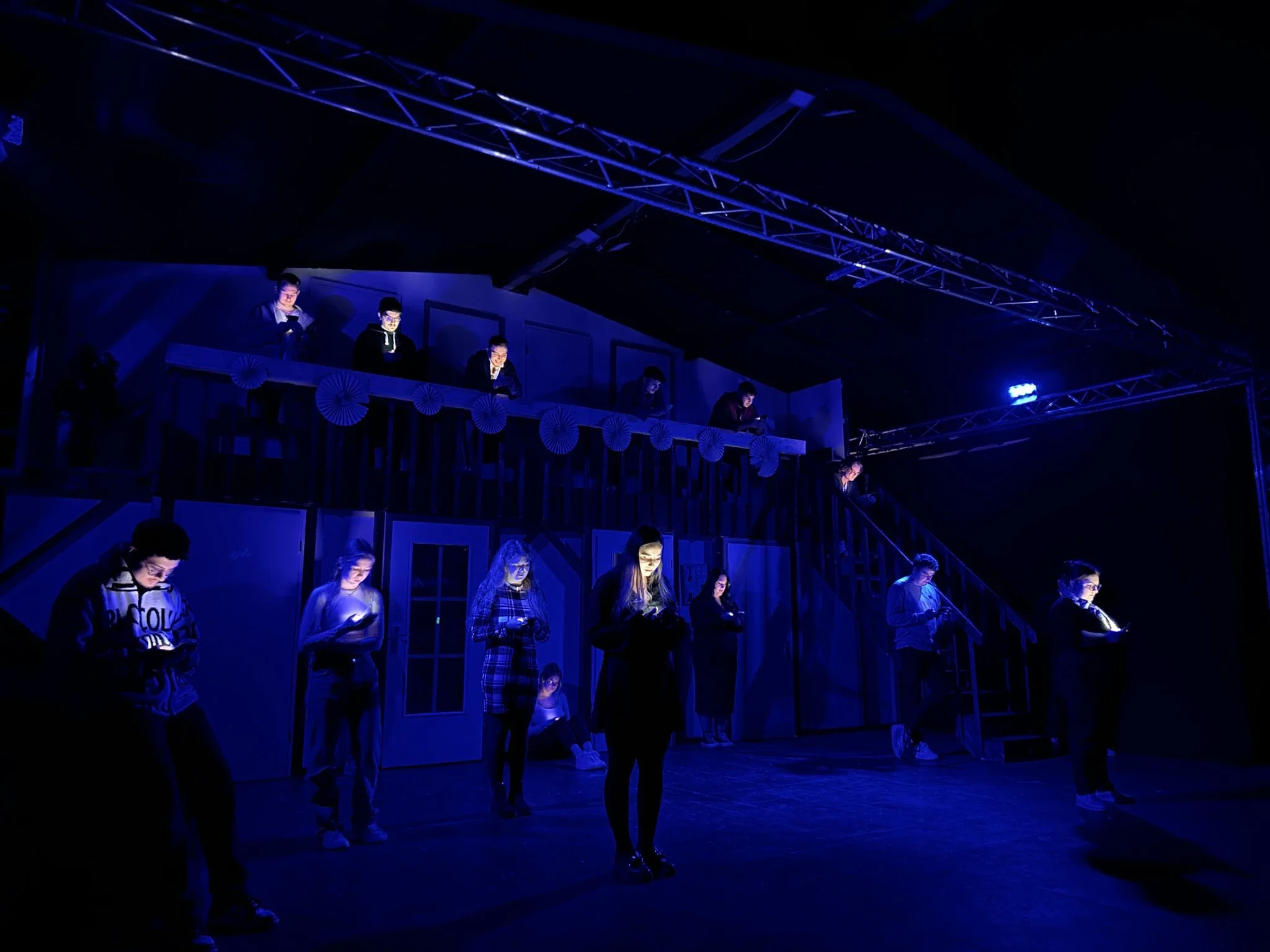 Bühne mit mehreren Schauspielern im Theater, beleuchtet in blauer Beleuchtung, einige stehen auf dem Boden, andere auf einer oberen Ebene, lesen Texte, kein Publikum sichtbar.