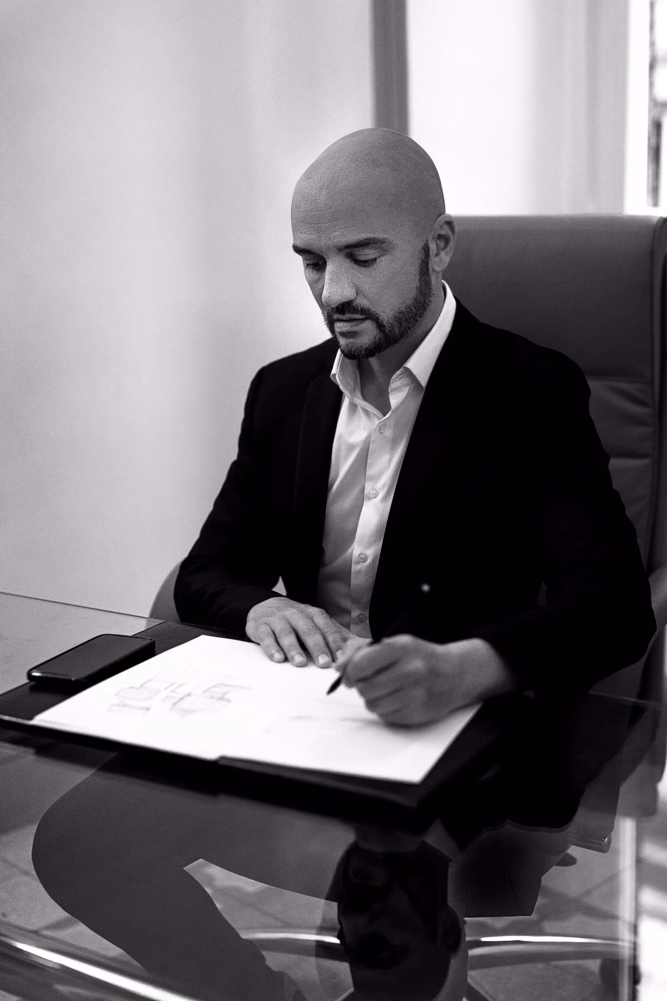 A man in a suit and white shirt sitting at a desk, looking down and writing on a large sheet of paper with a pen. There is a smartphone on the desk. The setting appears to be an office with plain walls and a window behind him.