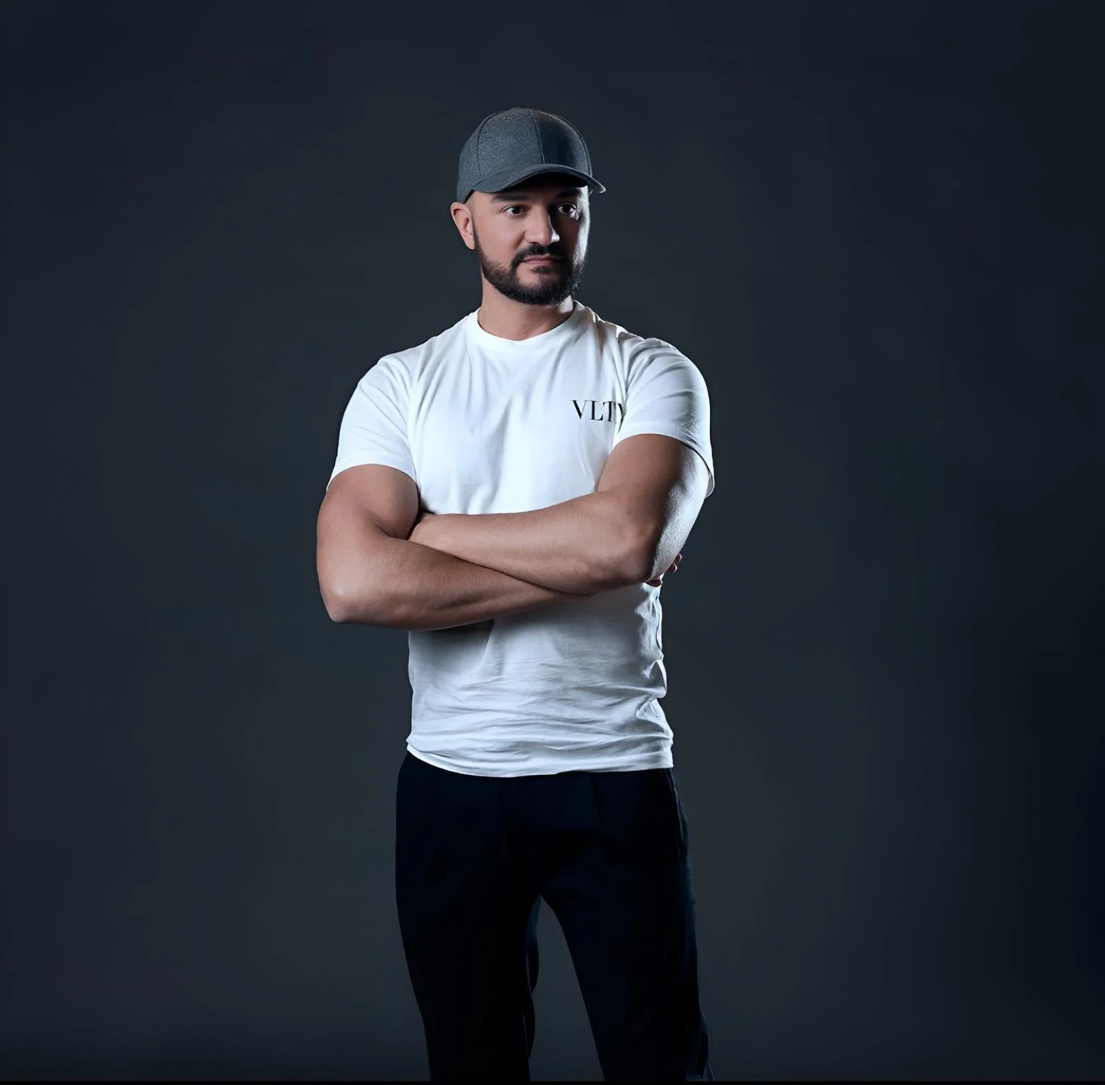 A young man with a beard wearing a gray cap, white T-shirt with 'VLTV' on it, and black pants, standing with arms crossed against a dark background.