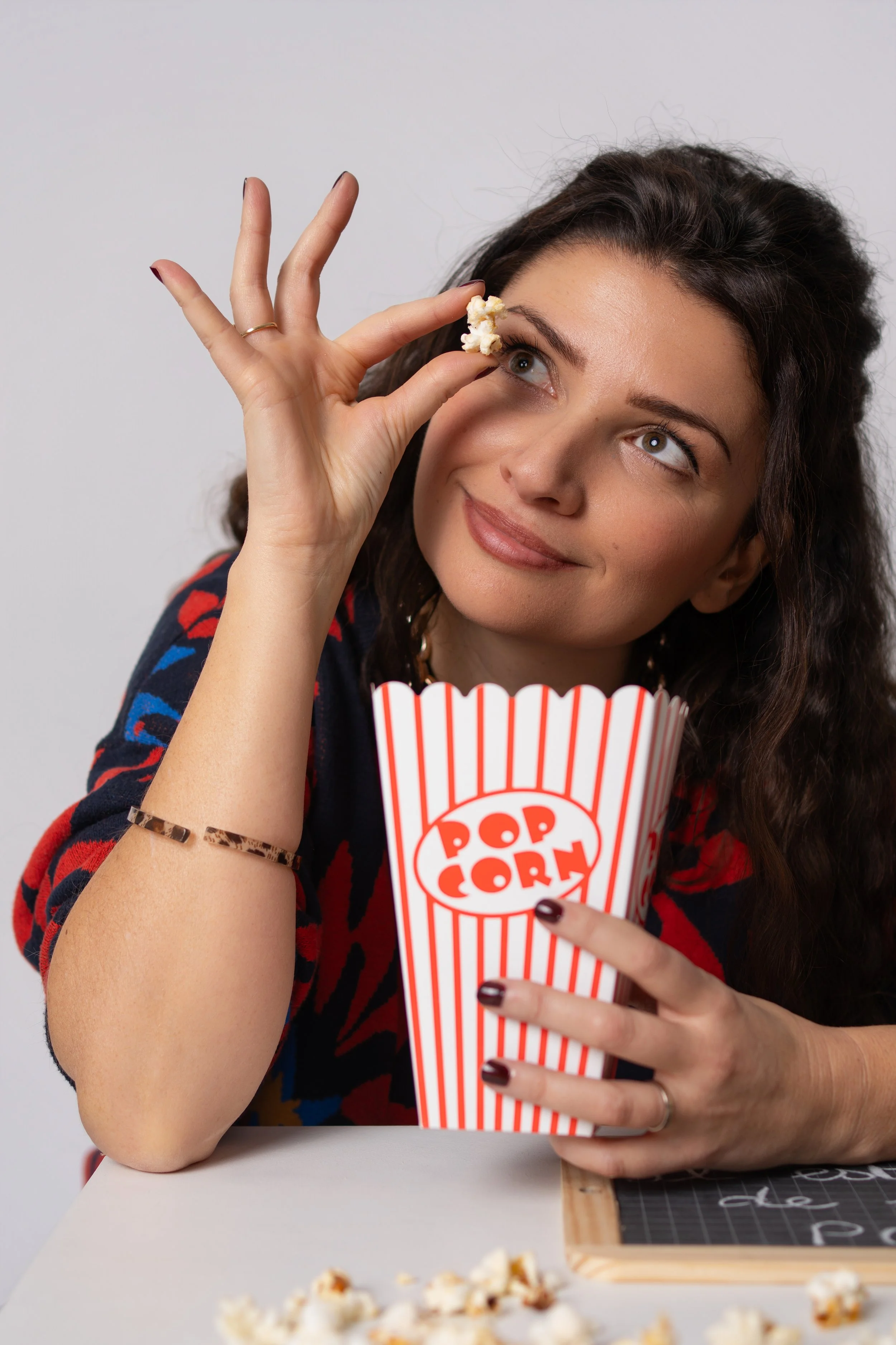 Femme à la peau claire, cheveux bruns ondulés, tenant un grand récipient à popcorn avec un sourire, portant un bracelet en lamelles et habillée d'une robe à motifs colorés, avec des éclats de popcorn sur la table et un tableau noir.