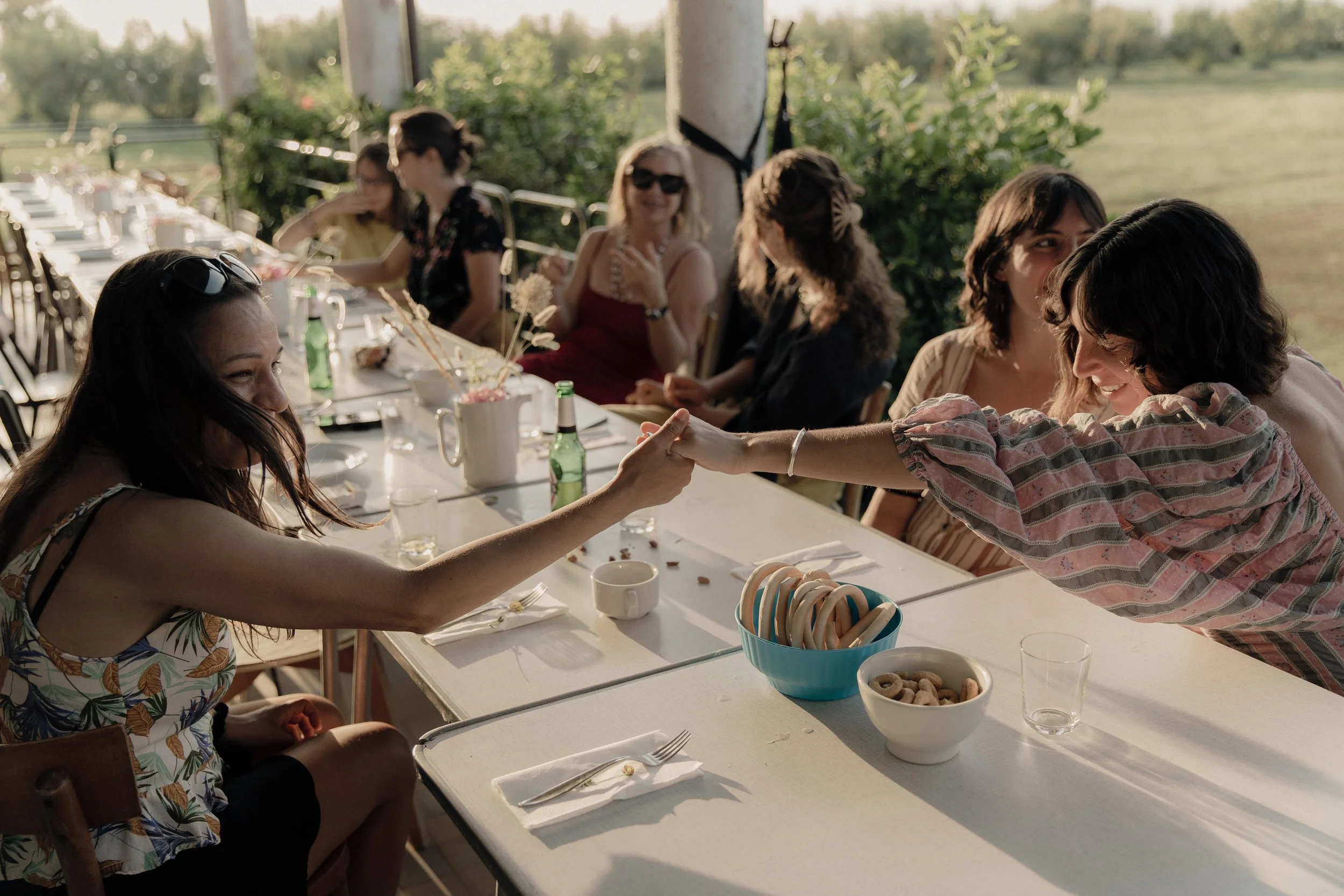 Un gruppo di donne sedute a un tavolo all'aperto in un ambiente estivo, condividendo un momento di festa con alcune in mano, mentre altre si sorridono e socializzano.