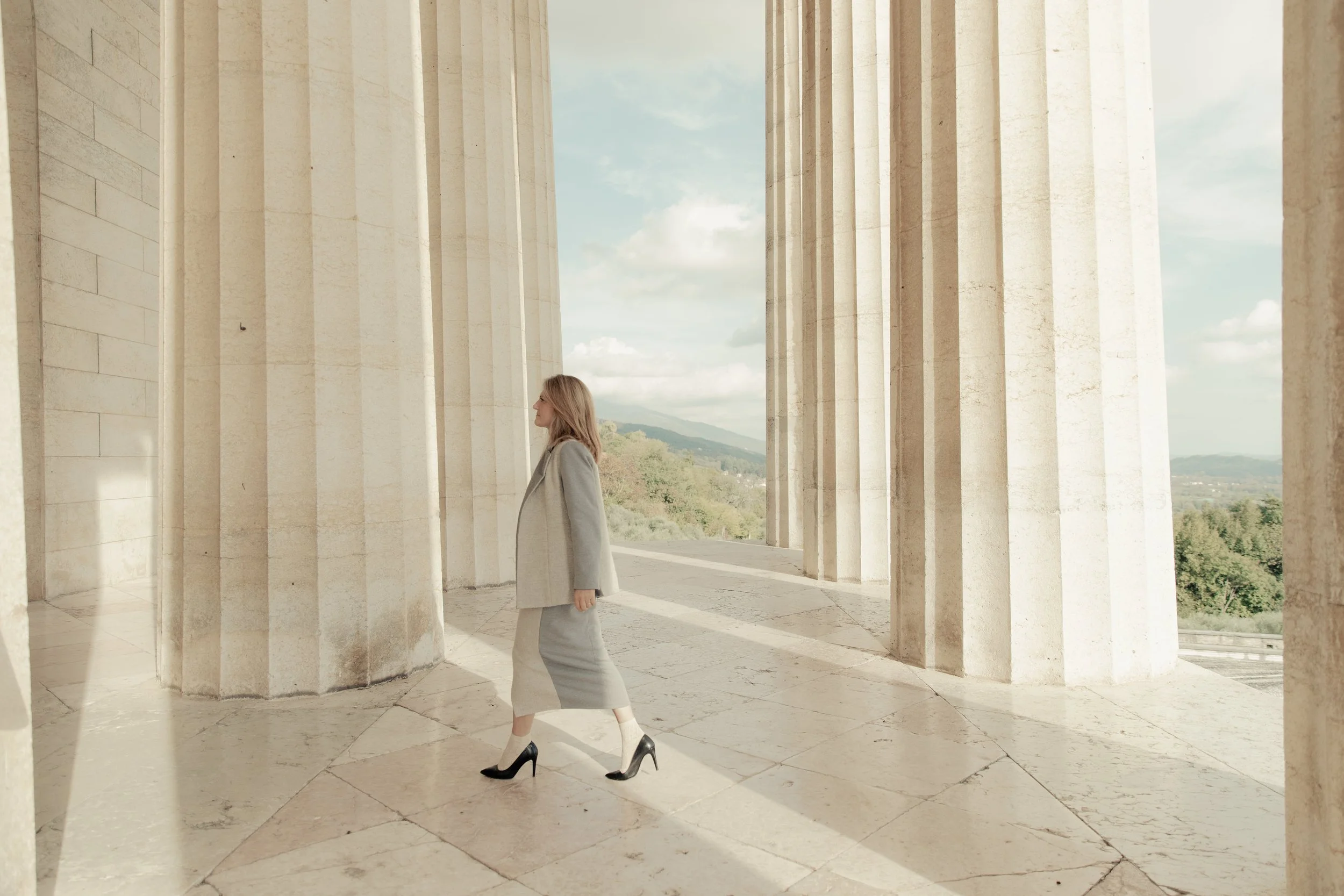 Donna in abbigliamento elegante cammina tra colonne di un edificio storico con vista panoramica sul paesaggio circostante.