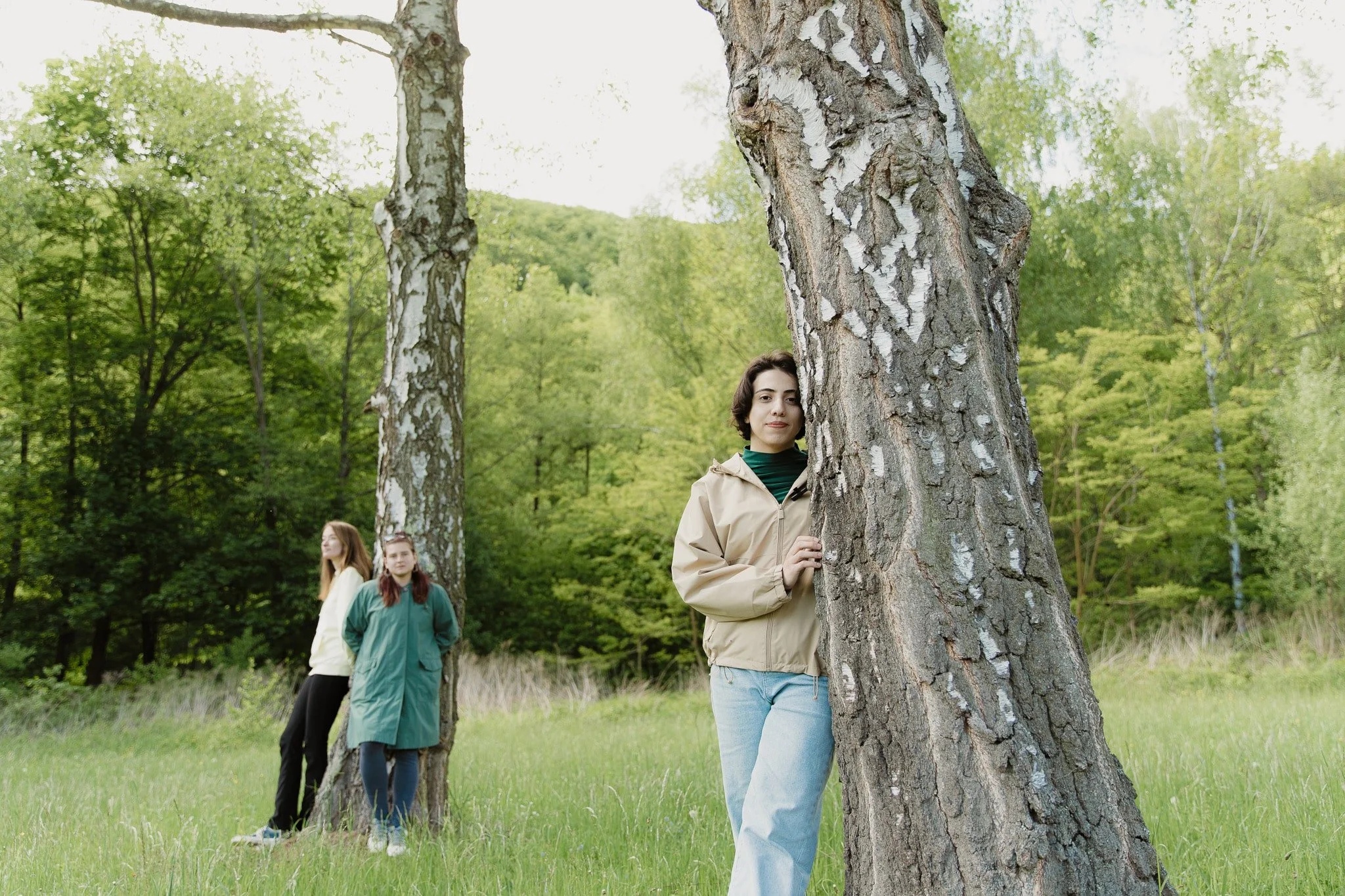Tre giovani donne in un prato di campagna circondate da alberi verdi, una si appoggia a un albero davanti e le altre due stanno in disparte, in un ambiente rilassante e naturale.