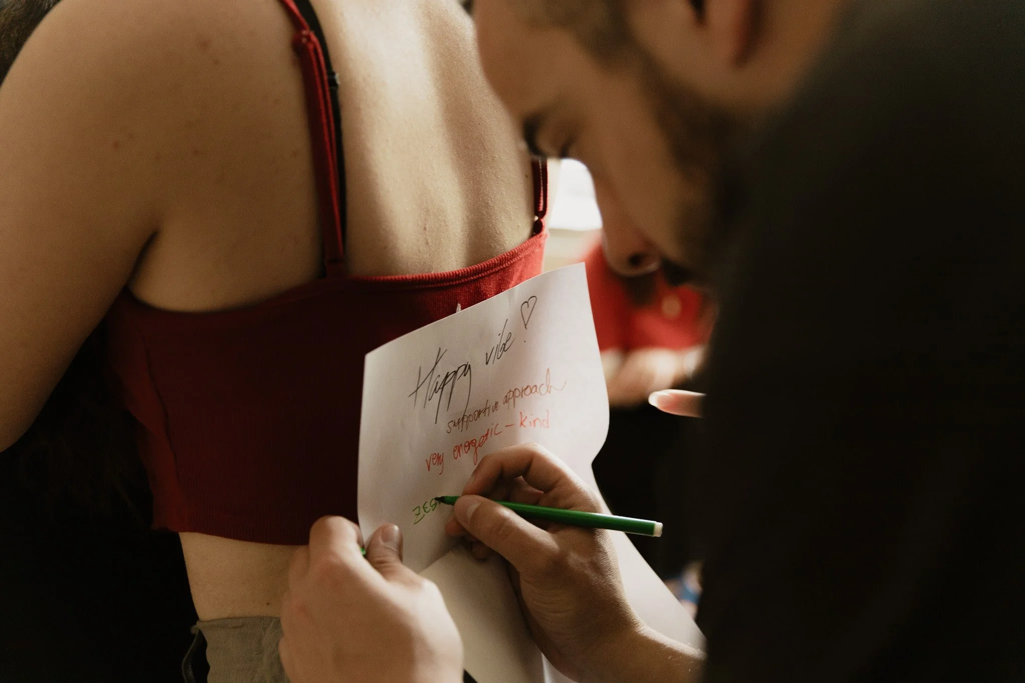 Una persona sta scrivendo un messaggio sulla schiena di un'altra persona con un pennarello verde, mentre questa tiene in mano un foglio con scritto 'Happy vade' e altre parole.