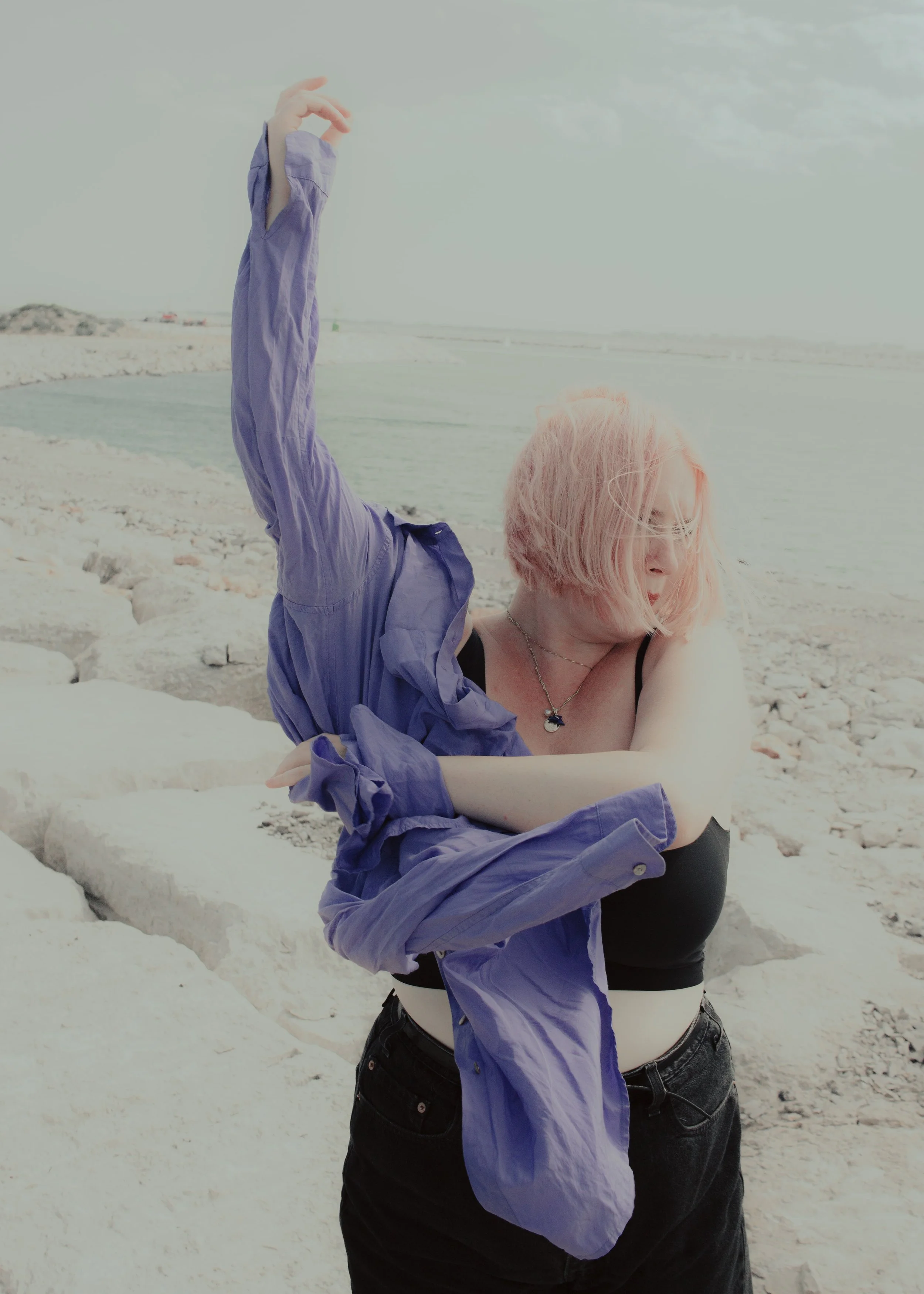 Una donna con capelli rosa chiaro si trova sulla spiaggia mentre si rimuove una camicia viola, indossa uno top nero e pantaloni neri, e si copre il volto con i capelli.