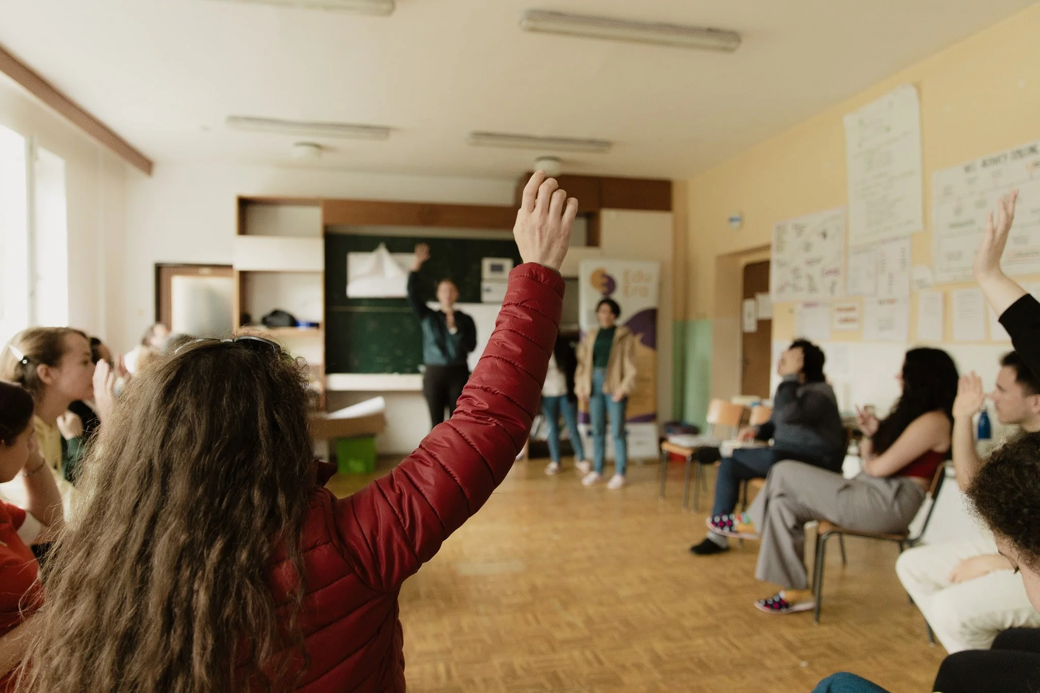 Classe scolastica con studenti che partecipano a una discussione, alcuni sollevano la mano, insegnanti alla lavagna, pareti decorate con disegni e appunti.