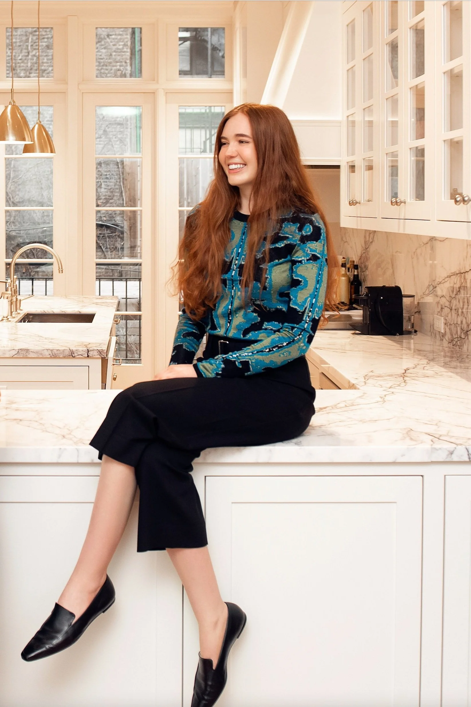 A woman with long red hair sitting on a marble kitchen countertop, wearing a patterned blouse, black pants, and black shoes, in a modern kitchen with white cabinets and large windows.