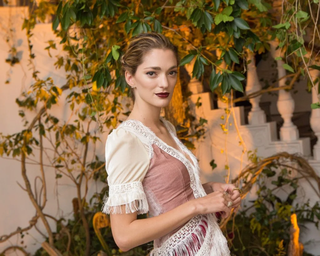 A woman posing indoors in front of greenery and a staircase, wearing a lace-trimmed outfit with short sleeves and a confident expression.