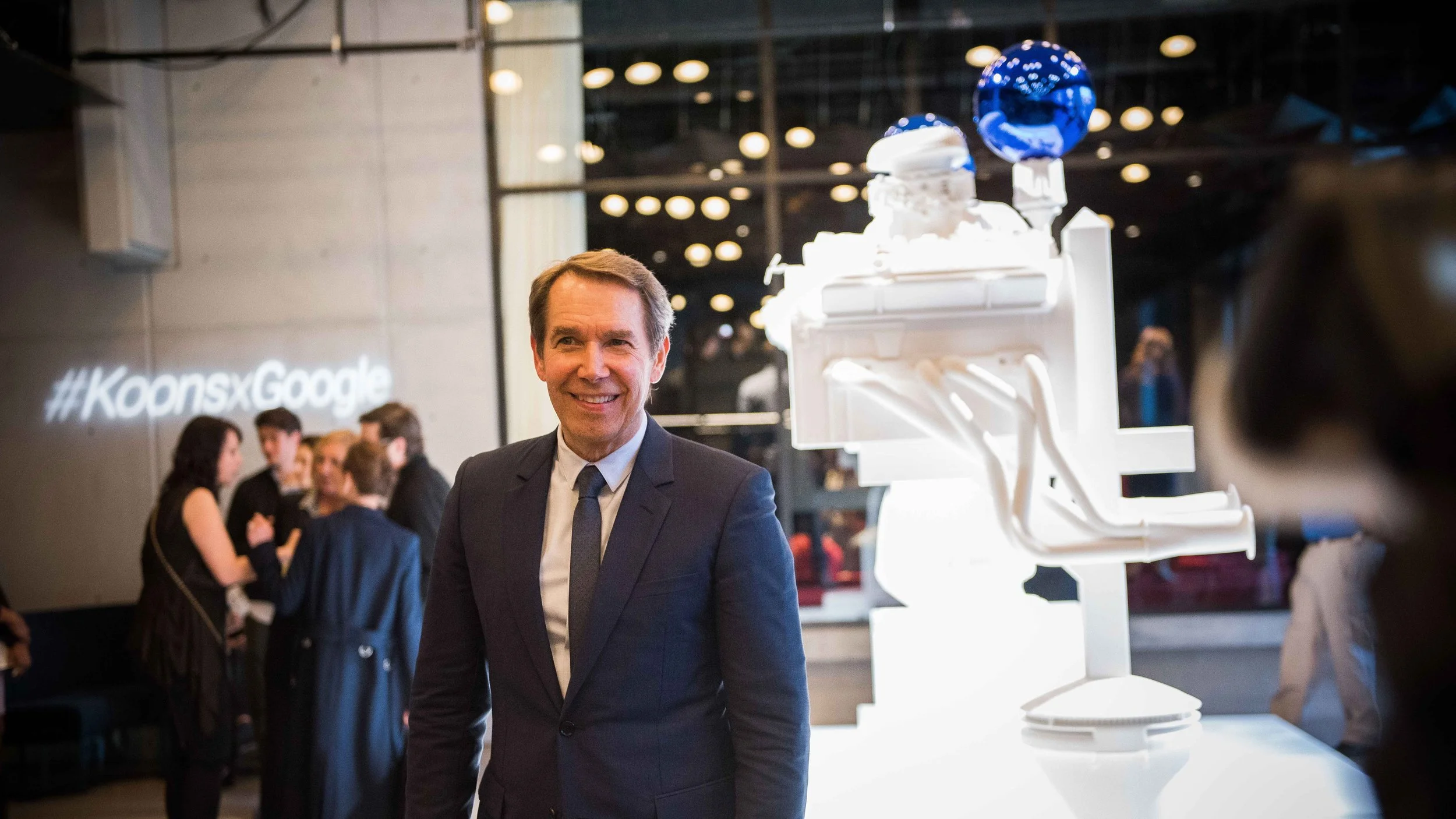 A man in a dark suit and tie smiling at a technology event with a white robot and blue globe sculpture behind him. In the background, several people are engaged in conversation, and the hashtag #KoonsxGoogle is projected on the wall.