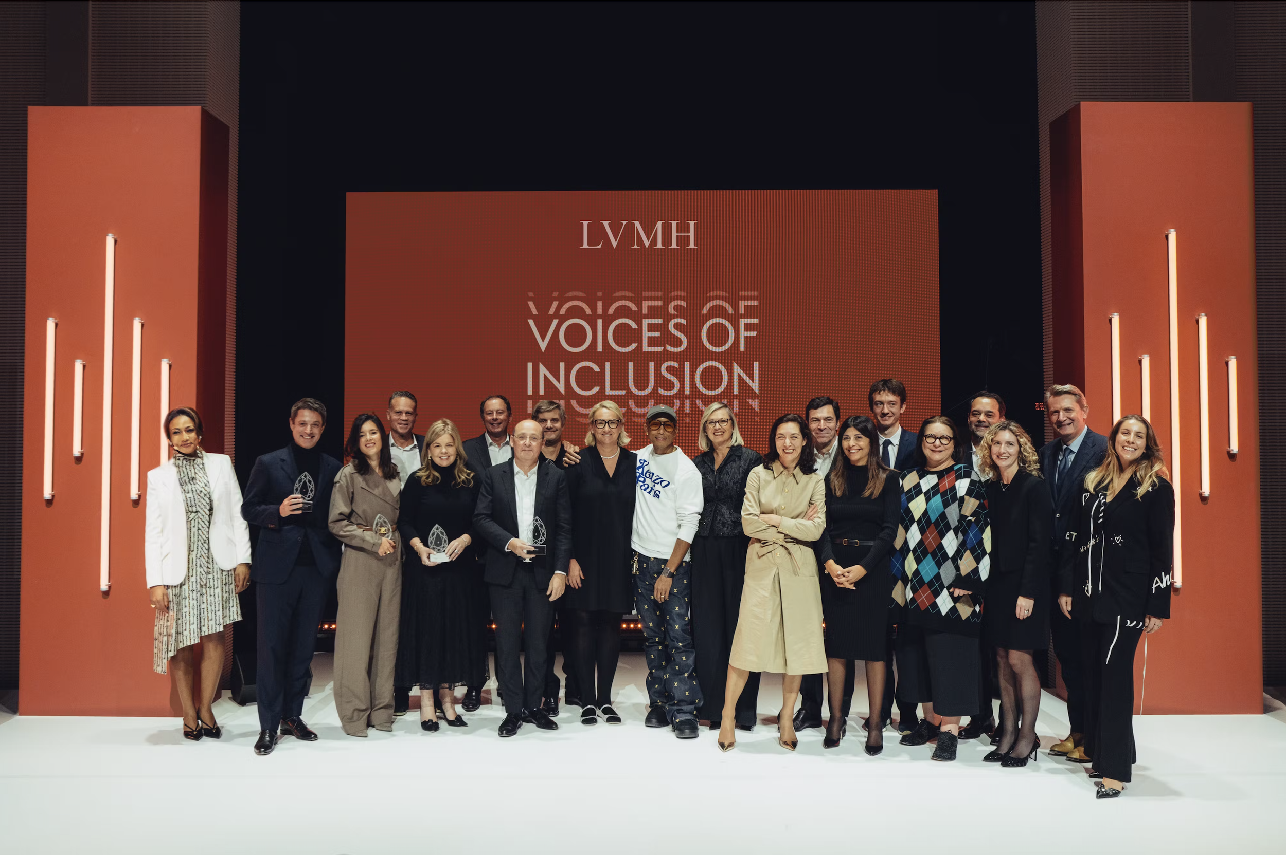 Group of people on stage at an event titled "Voices of Inclusion," with a large screen behind them displaying the event title and the LVMH logo, dressed in formal and semi-formal attire, holding awards, smiling for a photo.