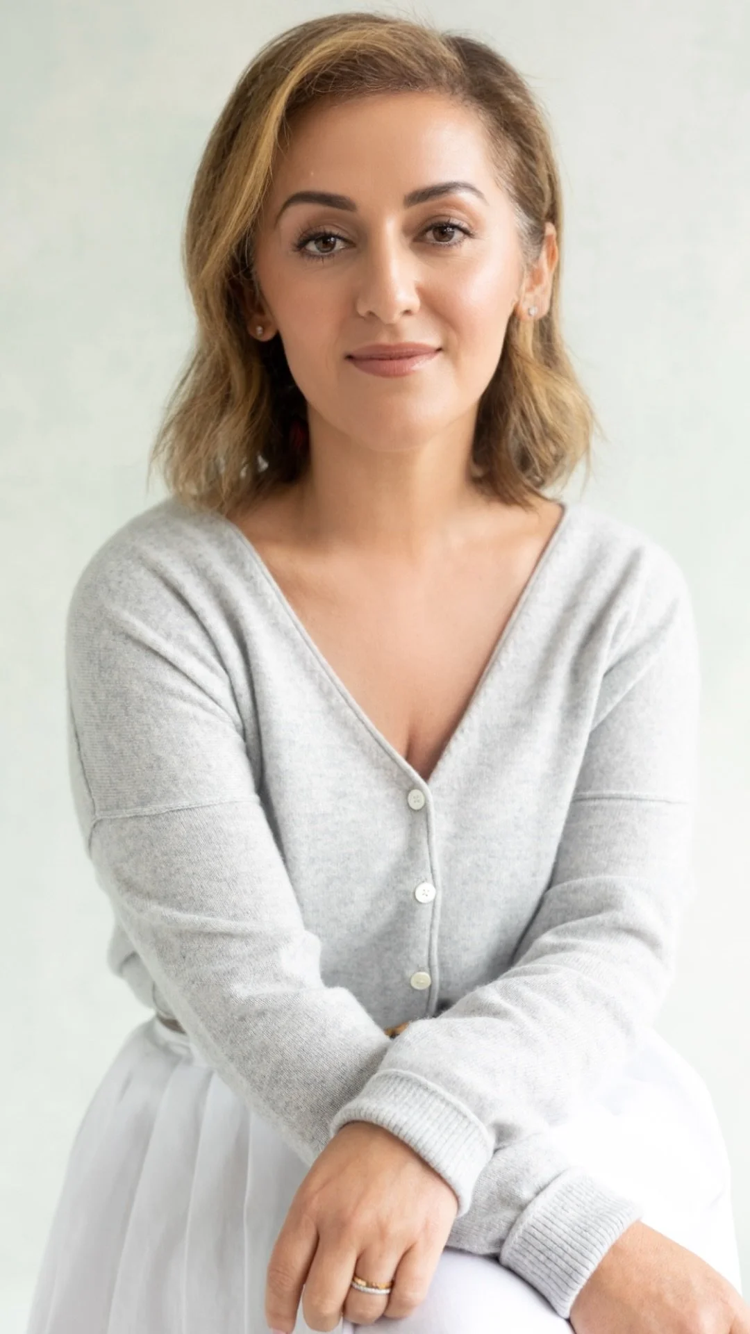 A woman with shoulder-length light brown hair and light skin, wearing a light gray button-up sweater and white skirt, sitting with crossed arms in front of a plain light background.