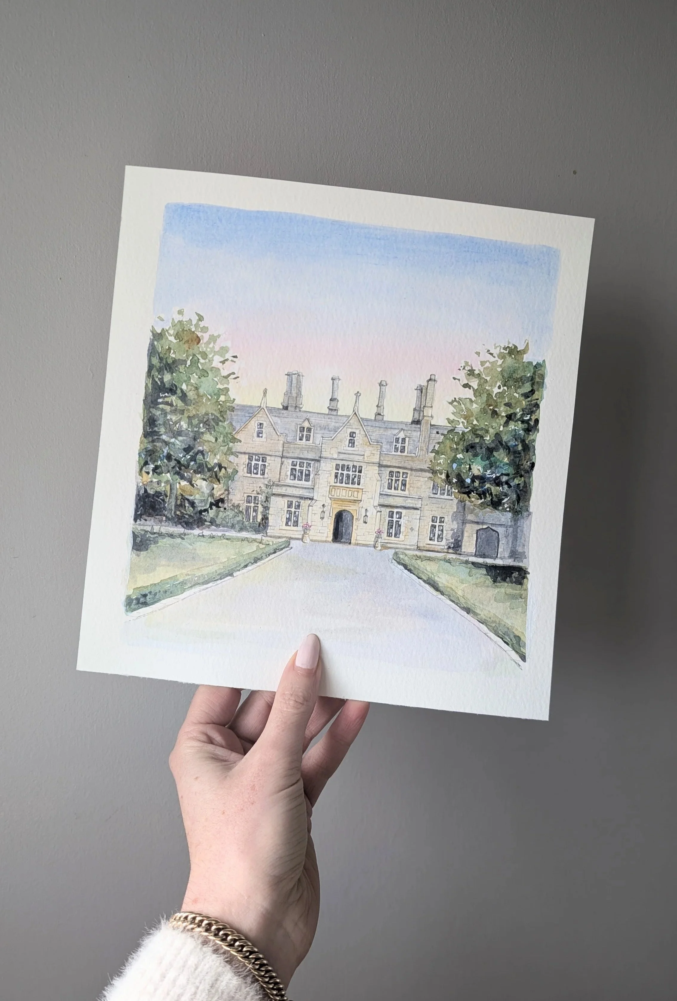 A hand holds a painting of Foxhills Golf club and wedding Venue in Ottershaw, Surrey.  Grand sandy coloured brick building with a soft sunset behind. A long driveway infront is flanked by grass and leafy trees. Painted by Kerry Regan. 