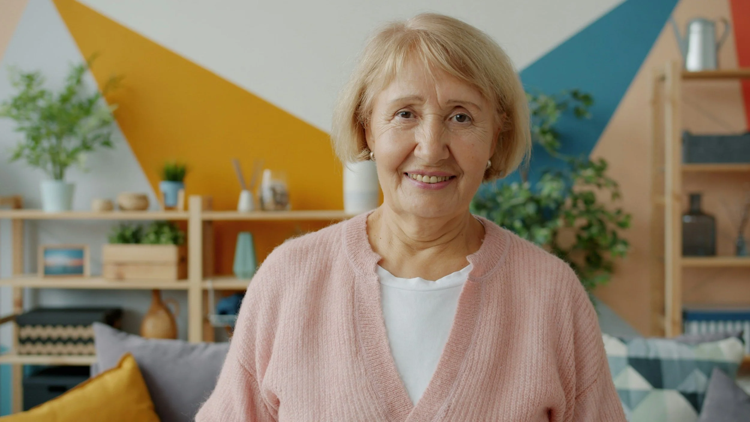 A smiling elderly woman with short blonde hair stands in a colorful, modern living room with plants and decorative objects on shelves in the background.
