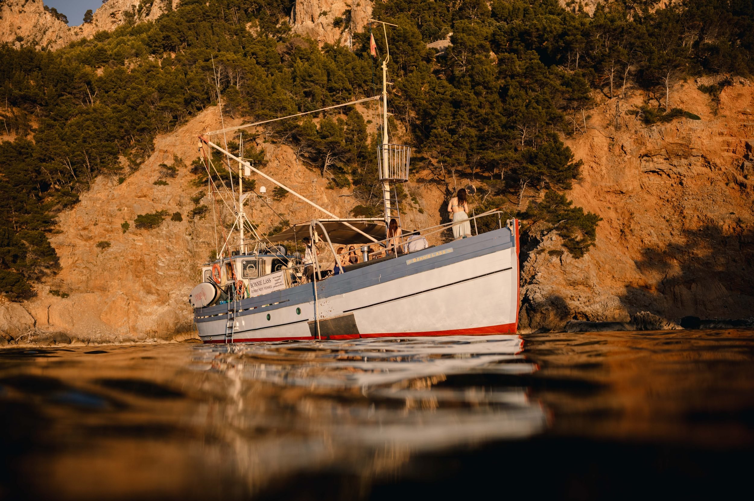 Bonnie Lass, a restored 1949 Scottish wooden fishing boat, anchored in a golden-lit cove on the Tramuntana coast