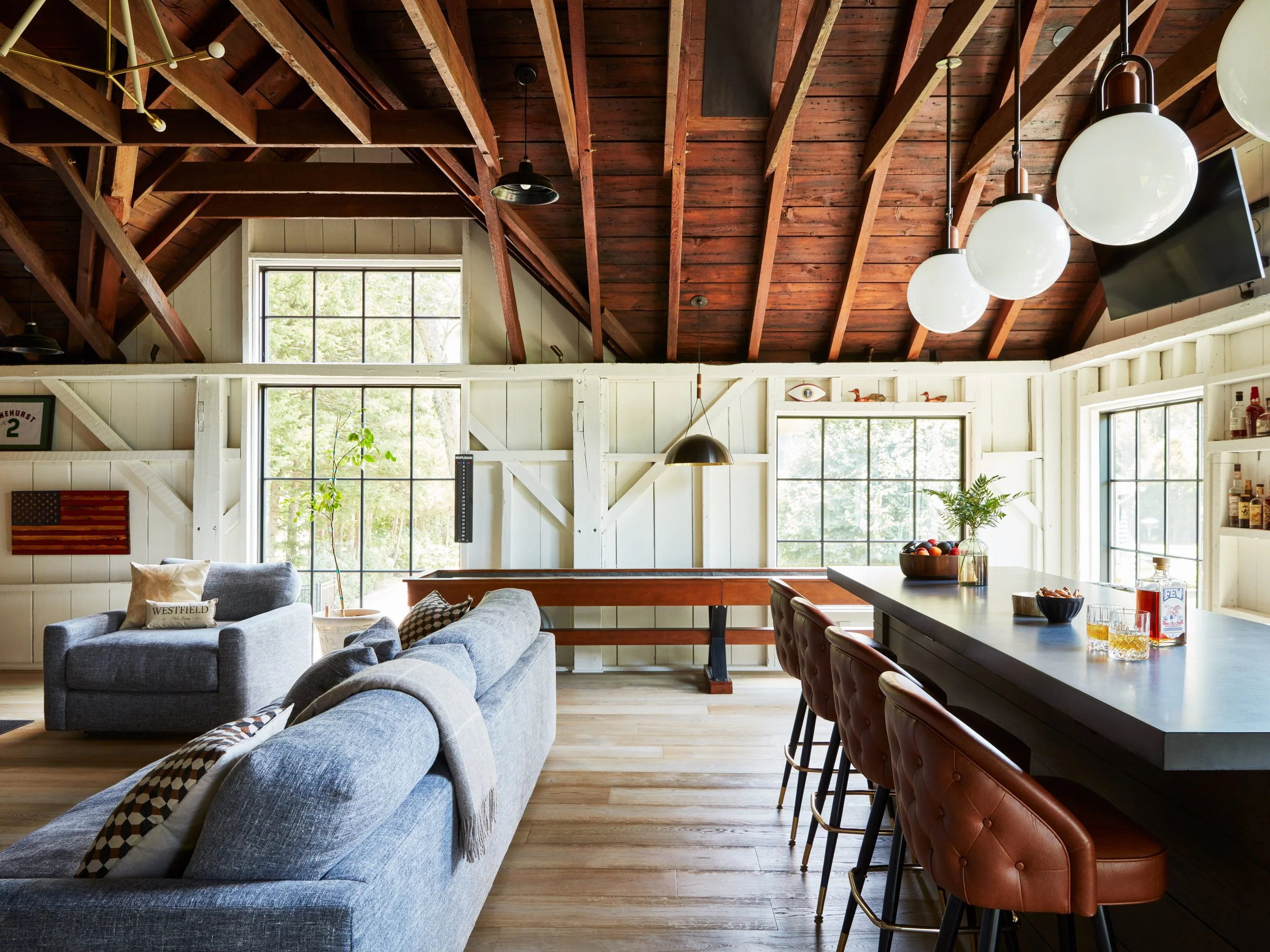 A spacious, bright living and dining area with large windows, wooden ceiling beams, and a mix of modern and rustic decor with sofas, bar stools, and hanging lights. Boutique hospitality recreation spatial and programming design.