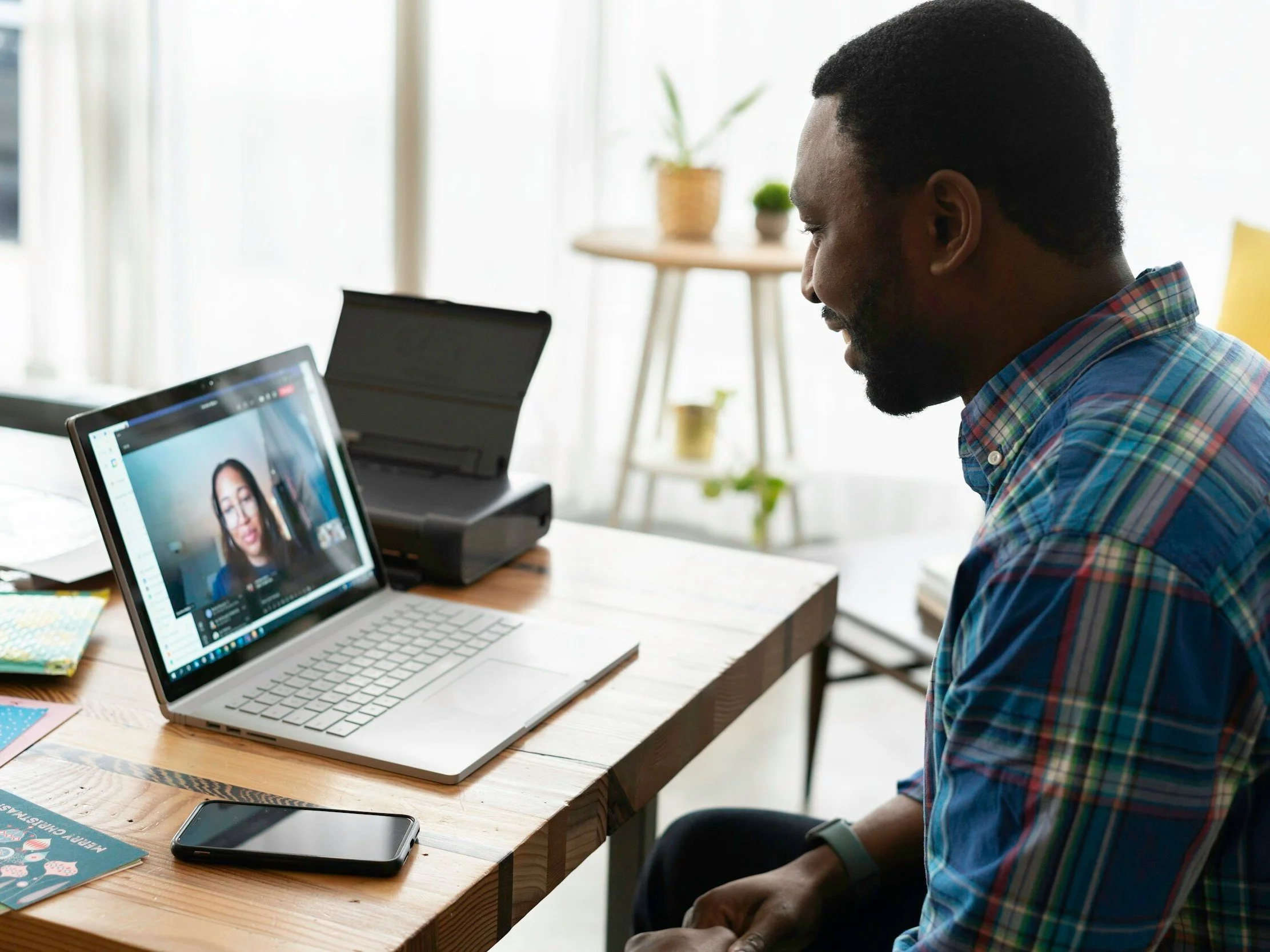 Man in a plaid shirt having a video call on his laptop in a bright room with plants and a wooden table.