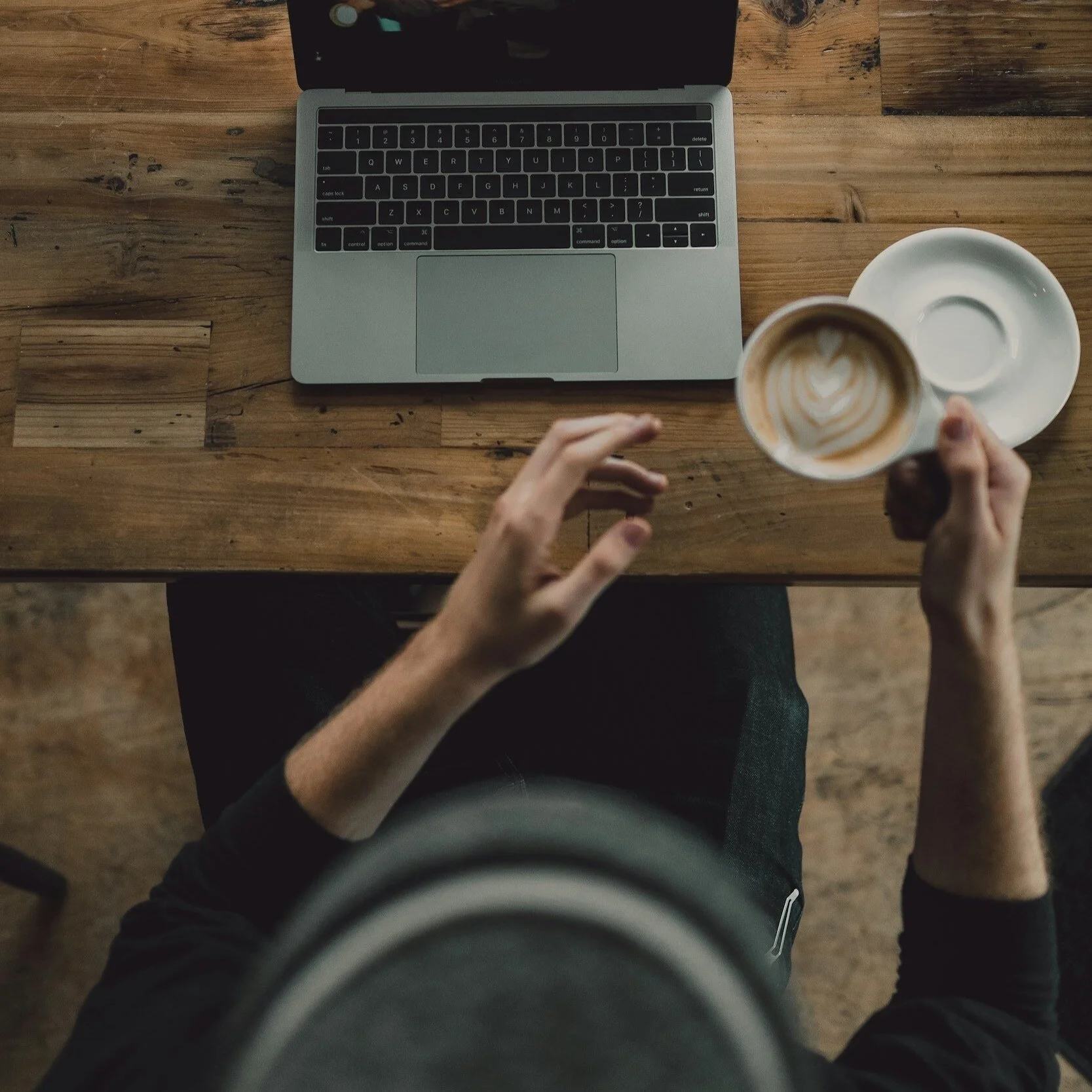 Overhead view of a person sitting at a wooden table with an open laptop, holding a cup of coffee with latte art, and an empty saucer nearby.