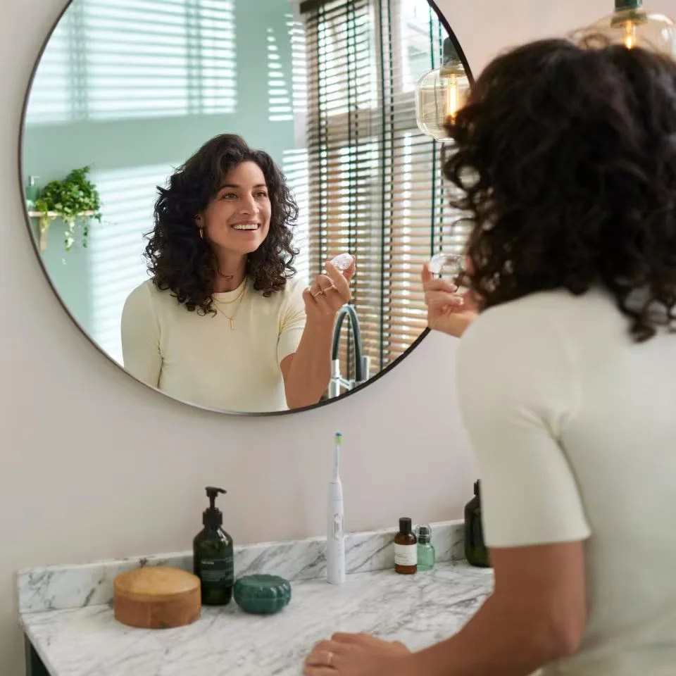 Une femme souriante se regarde dans un miroir en utilisant un dispositif dentaire électrique. La salle de bains a un plan de marbre et des bouteilles de produits de soins, avec des volets en bois et une plante verte en arrière-plan.