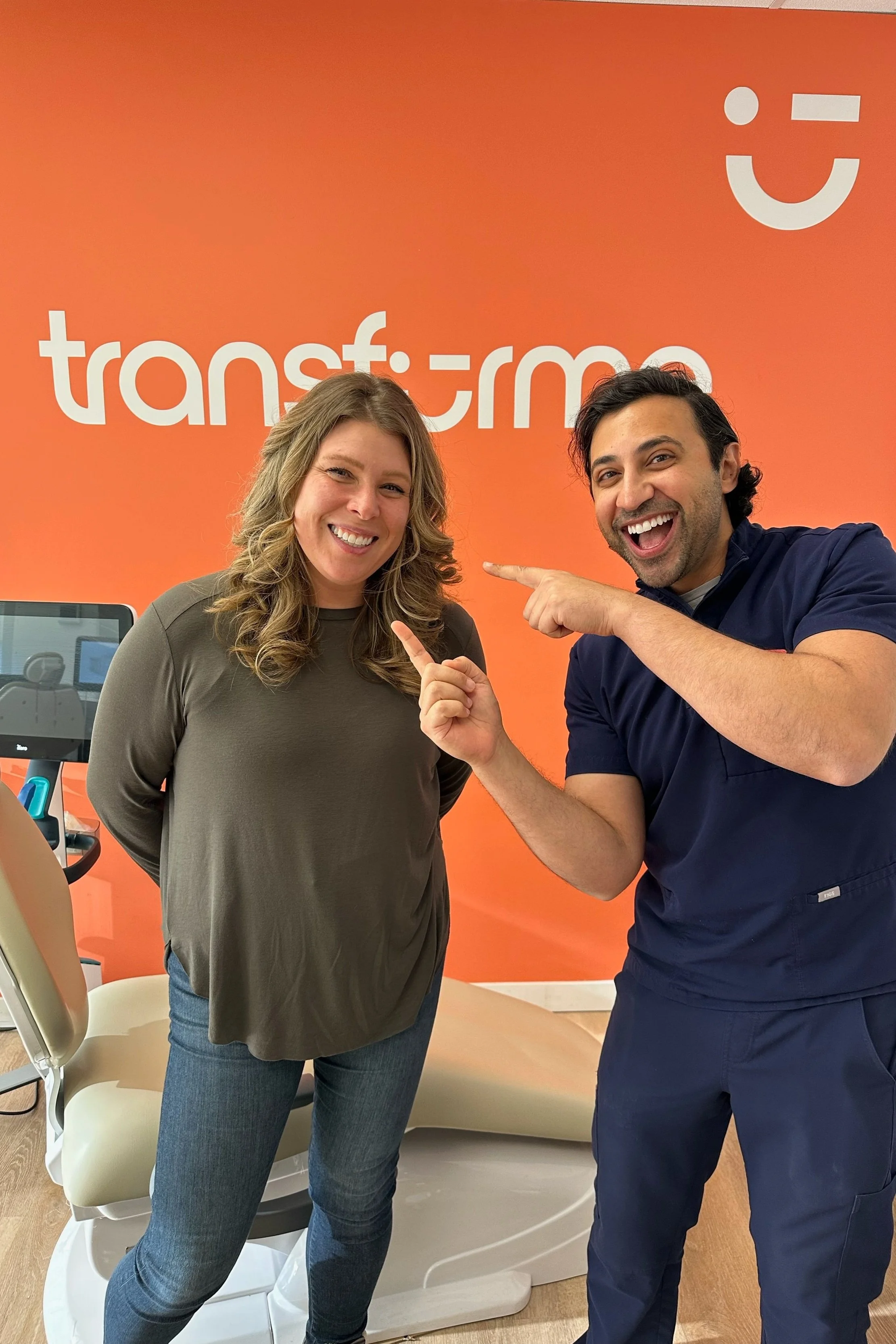 Deux personnes souriantes, un homme en vêtements médicaux et une femme en vêtements décontractés, posent devant un fond orange avec le logo et le nom 'docs-transfer'. L'homme pointe la femme avec enthousiasme.