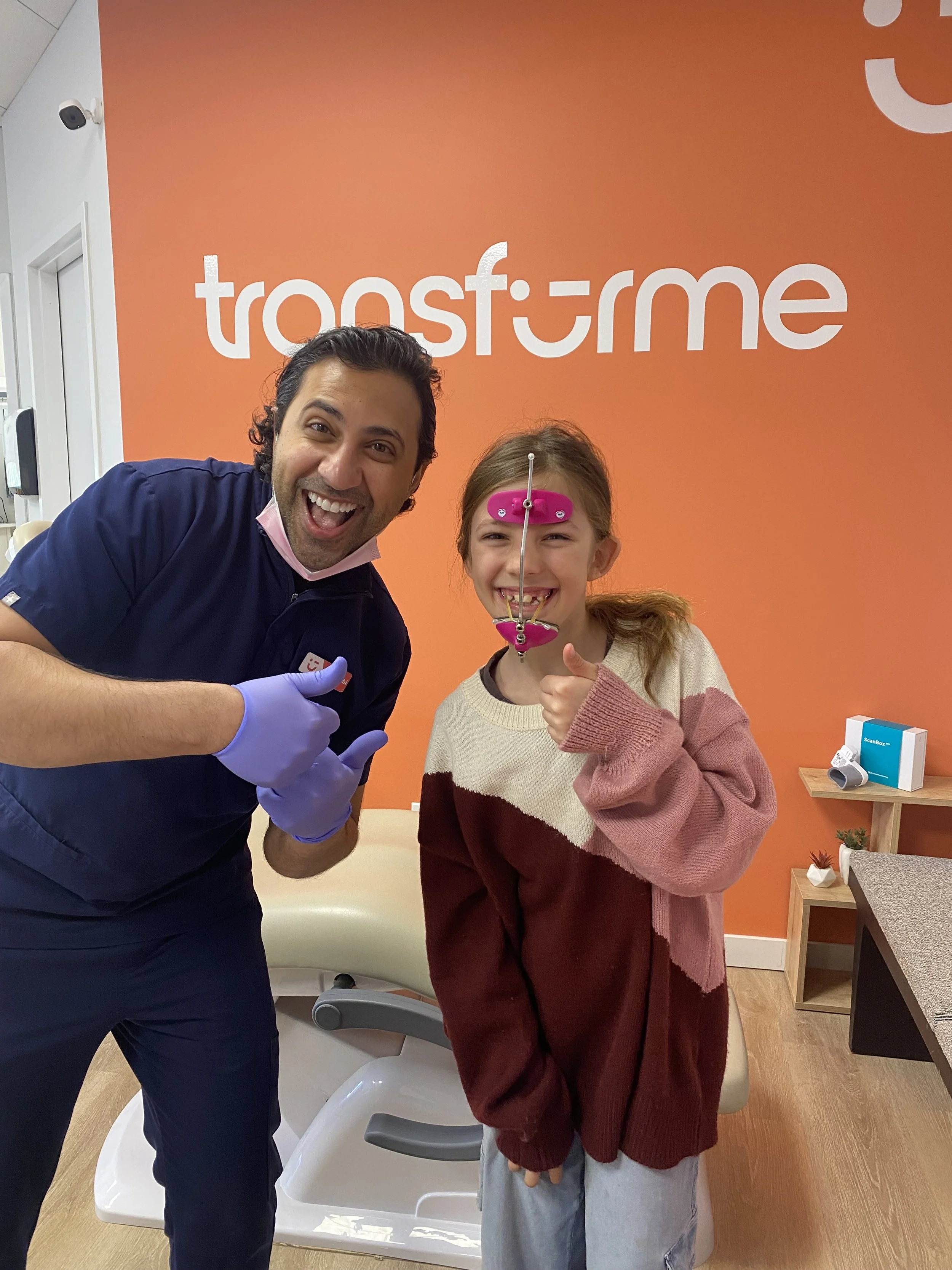 Une jeune fille et un professionnel de la santé souriant dans un centre médical, avec la jeune fille portant un électrodesur le visage, devant un mur orange avec le logo 'transforme'.