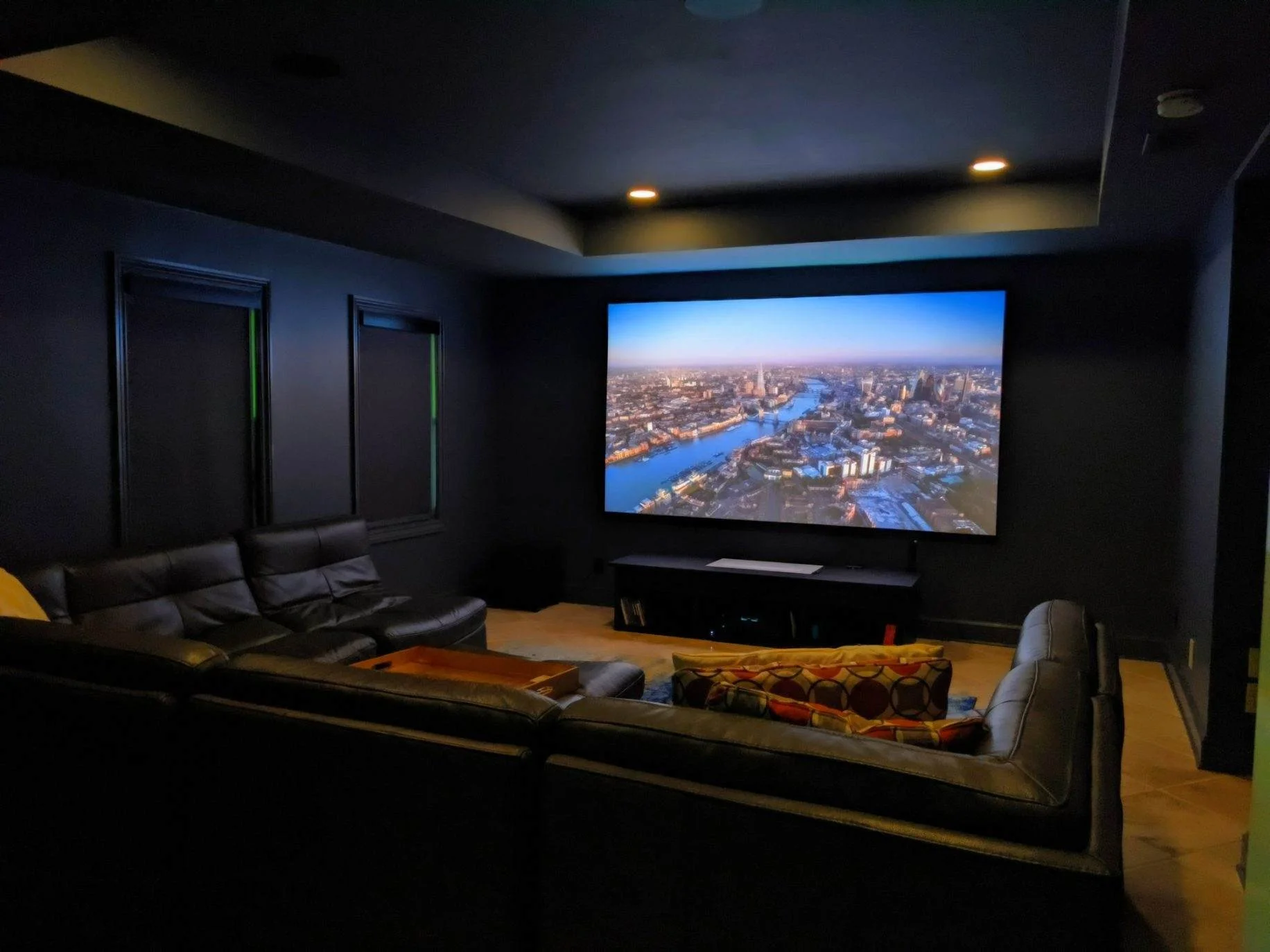 Home theater room with a large screen displaying a cityscape, black leather couch, and dark walls.