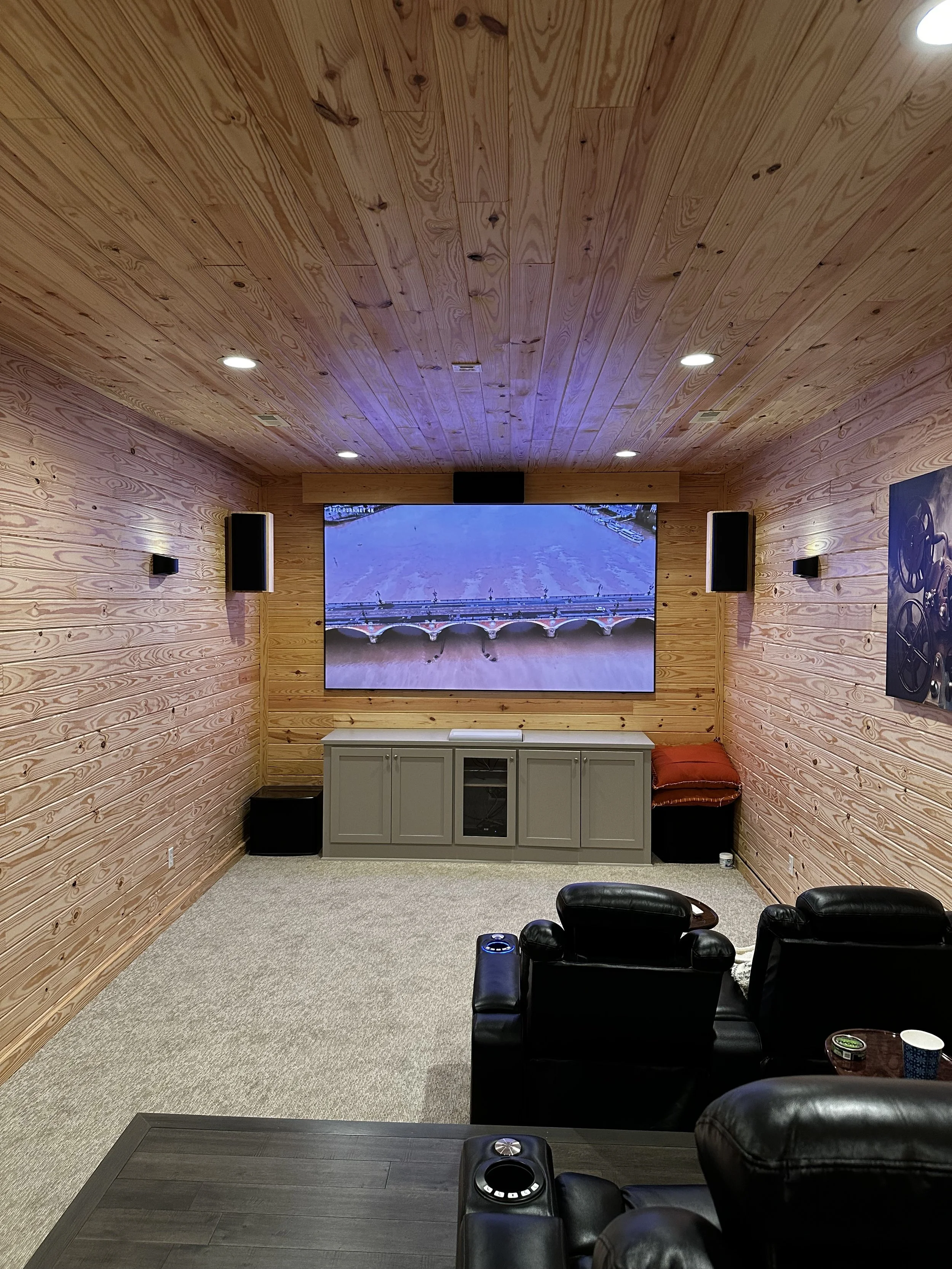 Home theater room with wood-paneled walls and ceiling, a large flat-screen TV showing a river scene, black reclining chairs, a gray cabinet beneath the TV, and wall-mounted speakers.