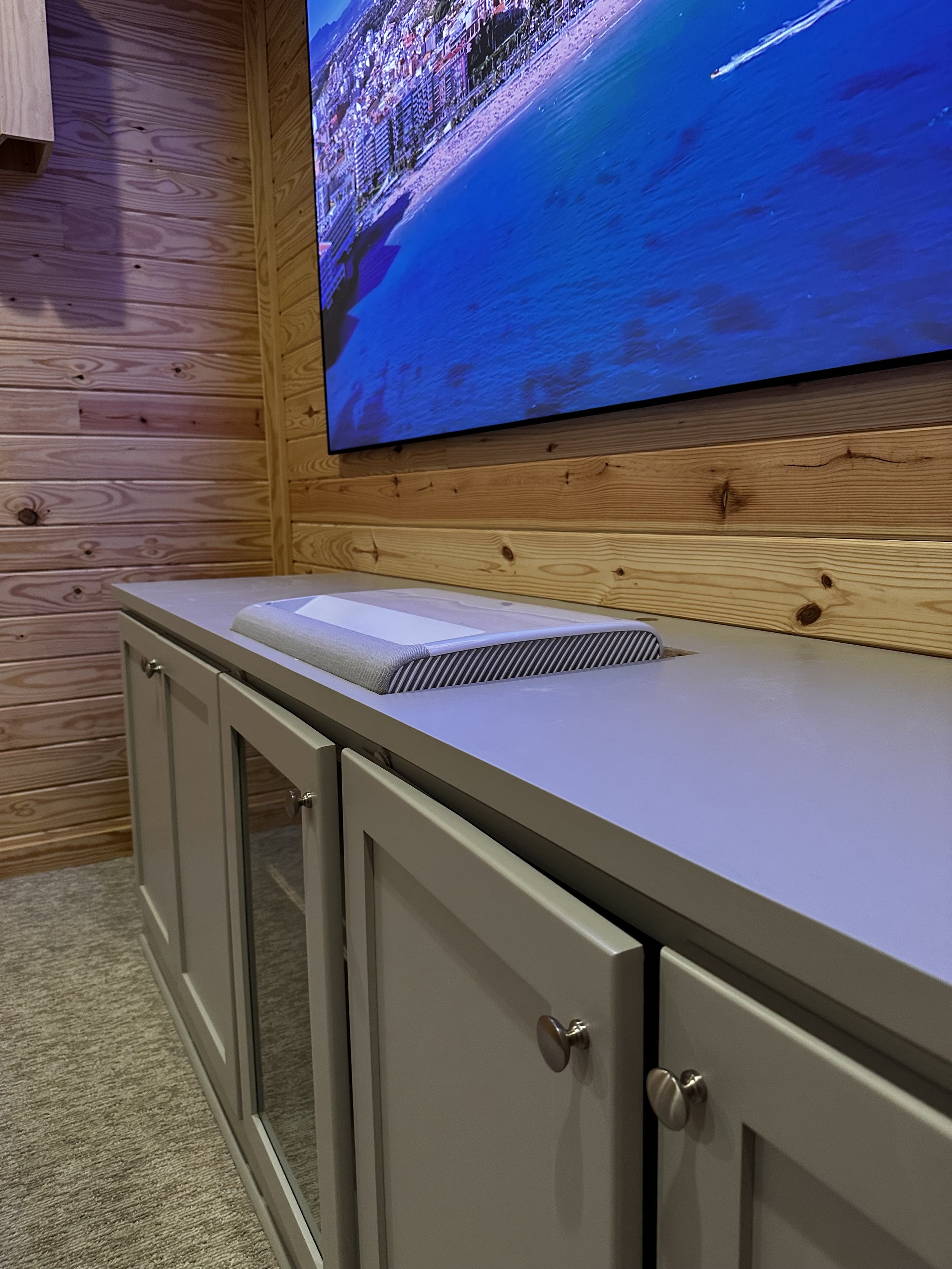 A wooden wall with a mounted flat-screen TV displaying an aerial view of a city coastline. Below the TV is a white cabinet with silver knobs and an object on top that is unclear.