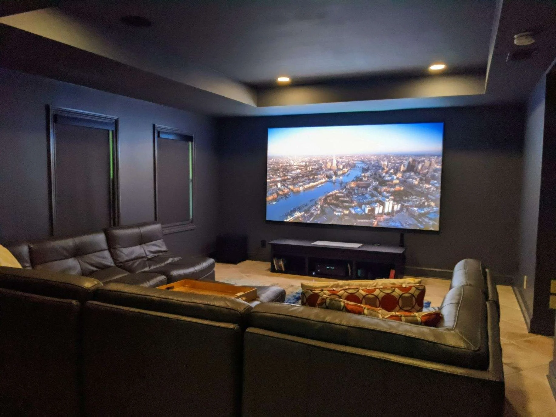 A home theater room with dark walls and ceiling, a large flat-screen TV displaying an aerial view of a city, a dark leather sectional sofa, a coffee table, and a media console.