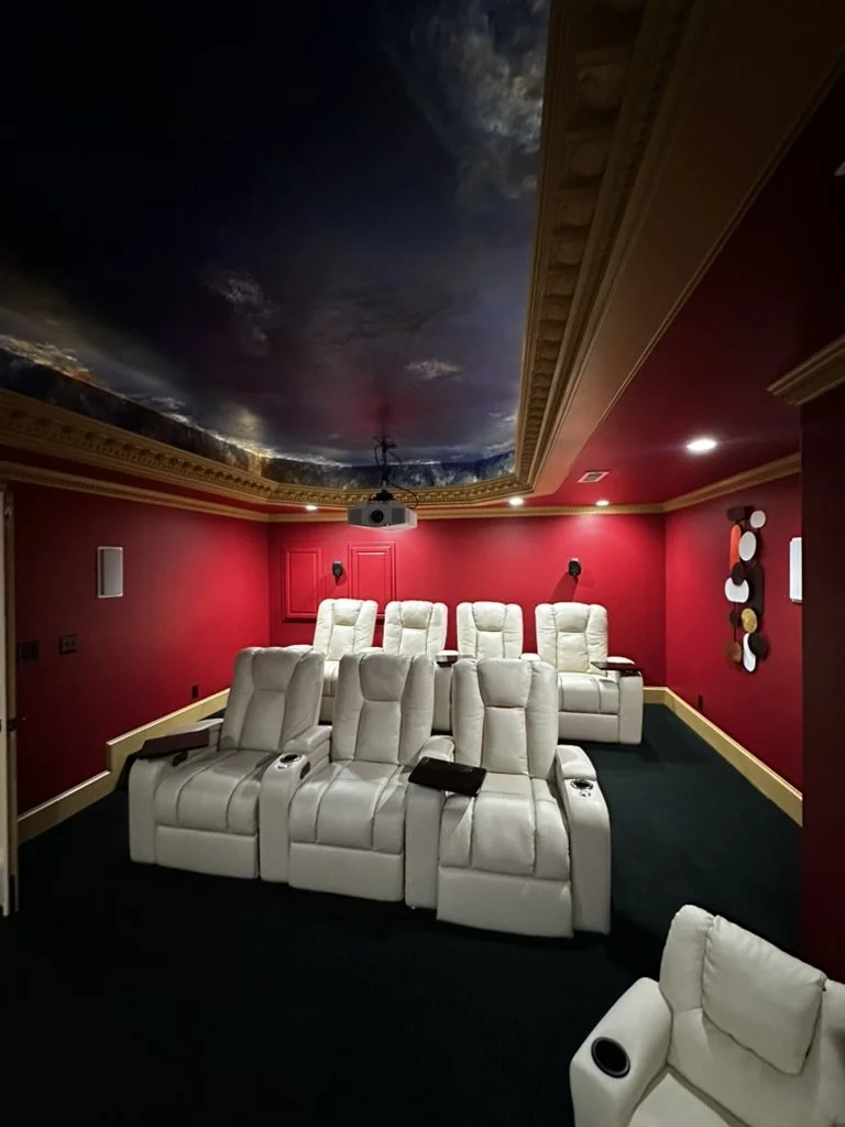 A home theater room with red walls, black carpet, and white reclining theater chairs. The ceiling has a sky mural with clouds and astronomy, and a projector is mounted on the ceiling. There are black wall-mounted speakers and colorful circular wall art.