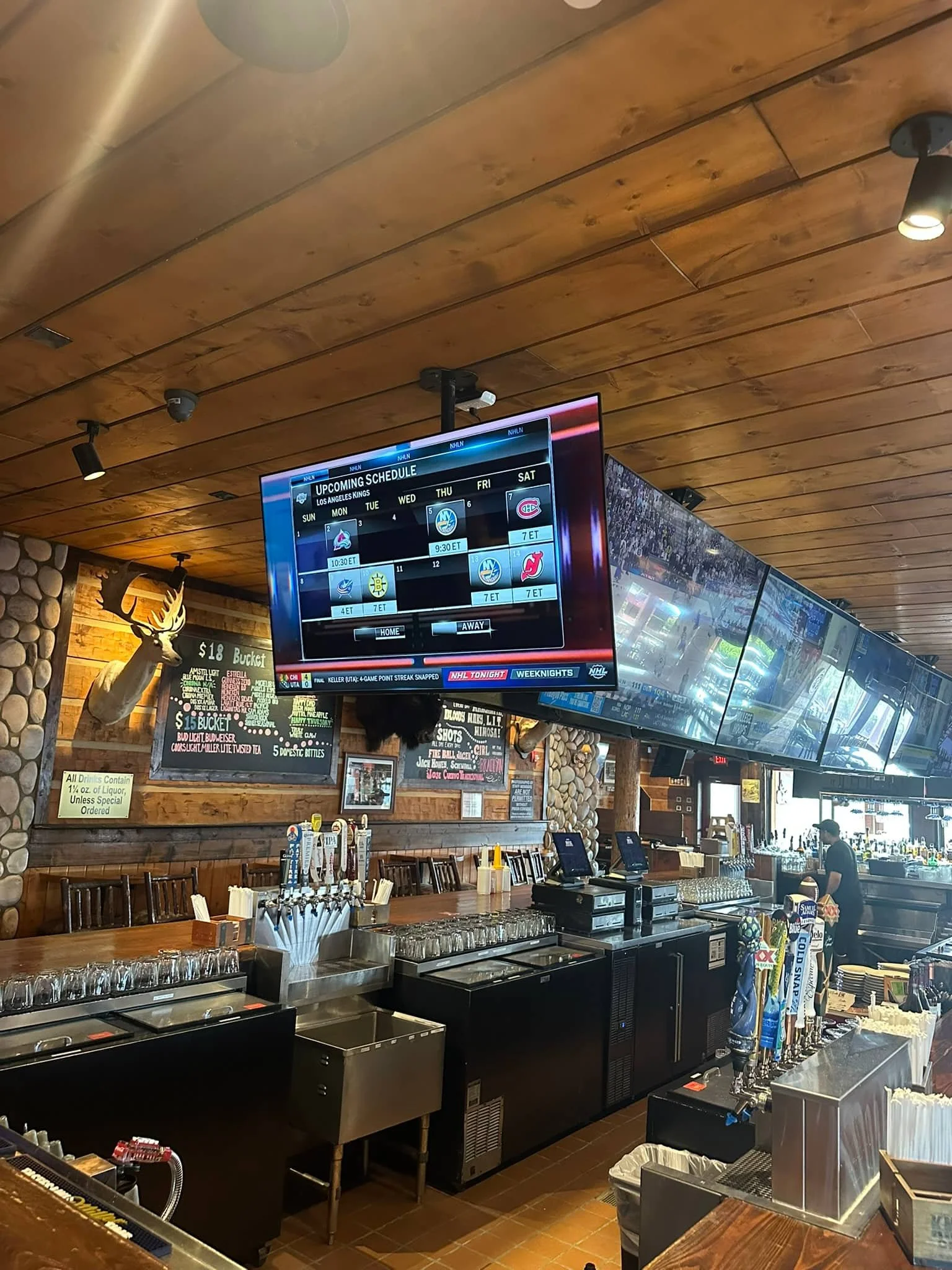 View of a bar or restaurant interior with a wooden ceiling, a TV displaying sports scores, and a bartender or server behind the counter. The bar area has beer taps, glasses, and some signs on the wall.
