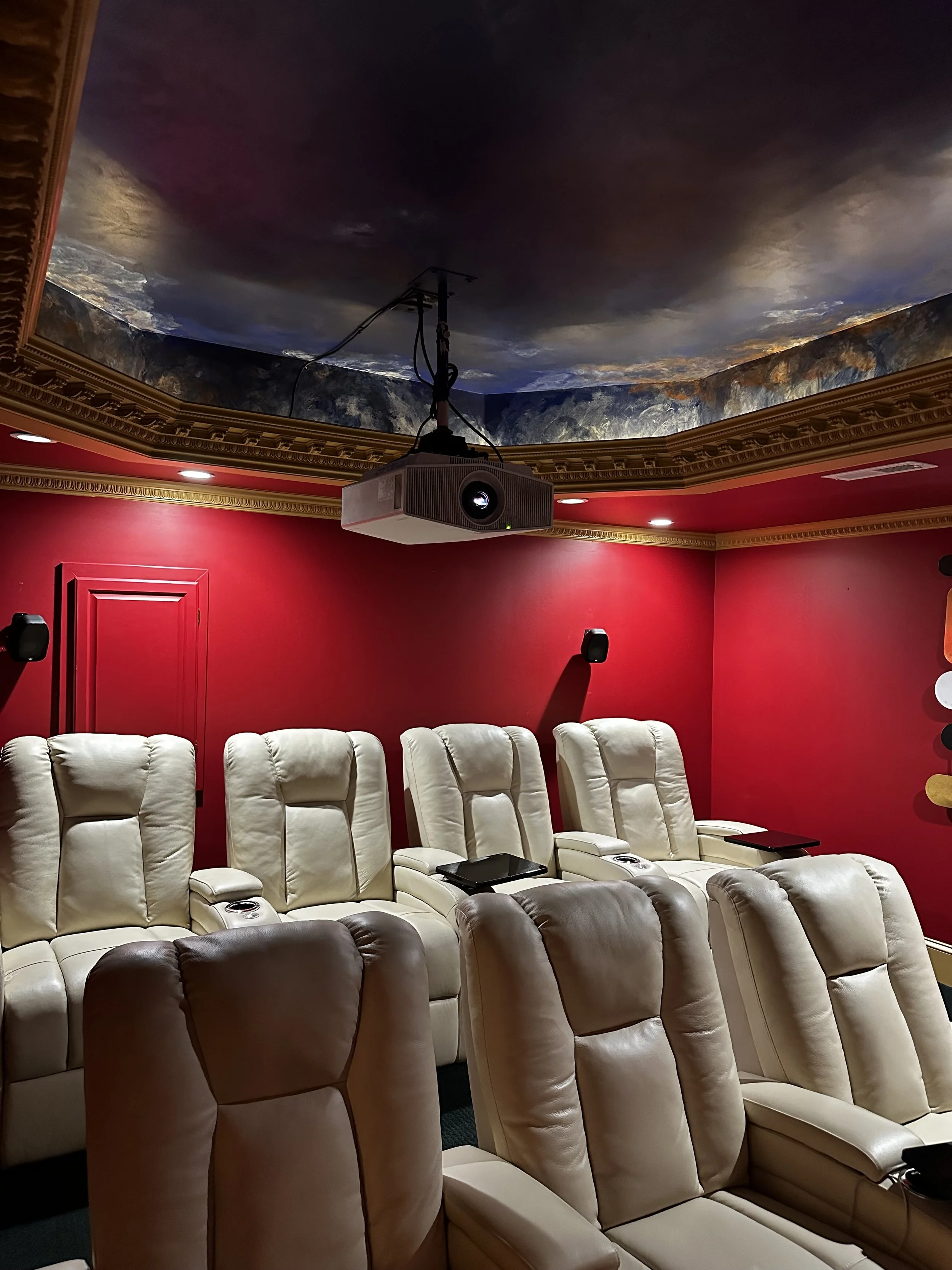 Home theater room with red walls, cream-colored reclining seats, and a projector mounted on the ceiling under a painted sky ceiling mural.