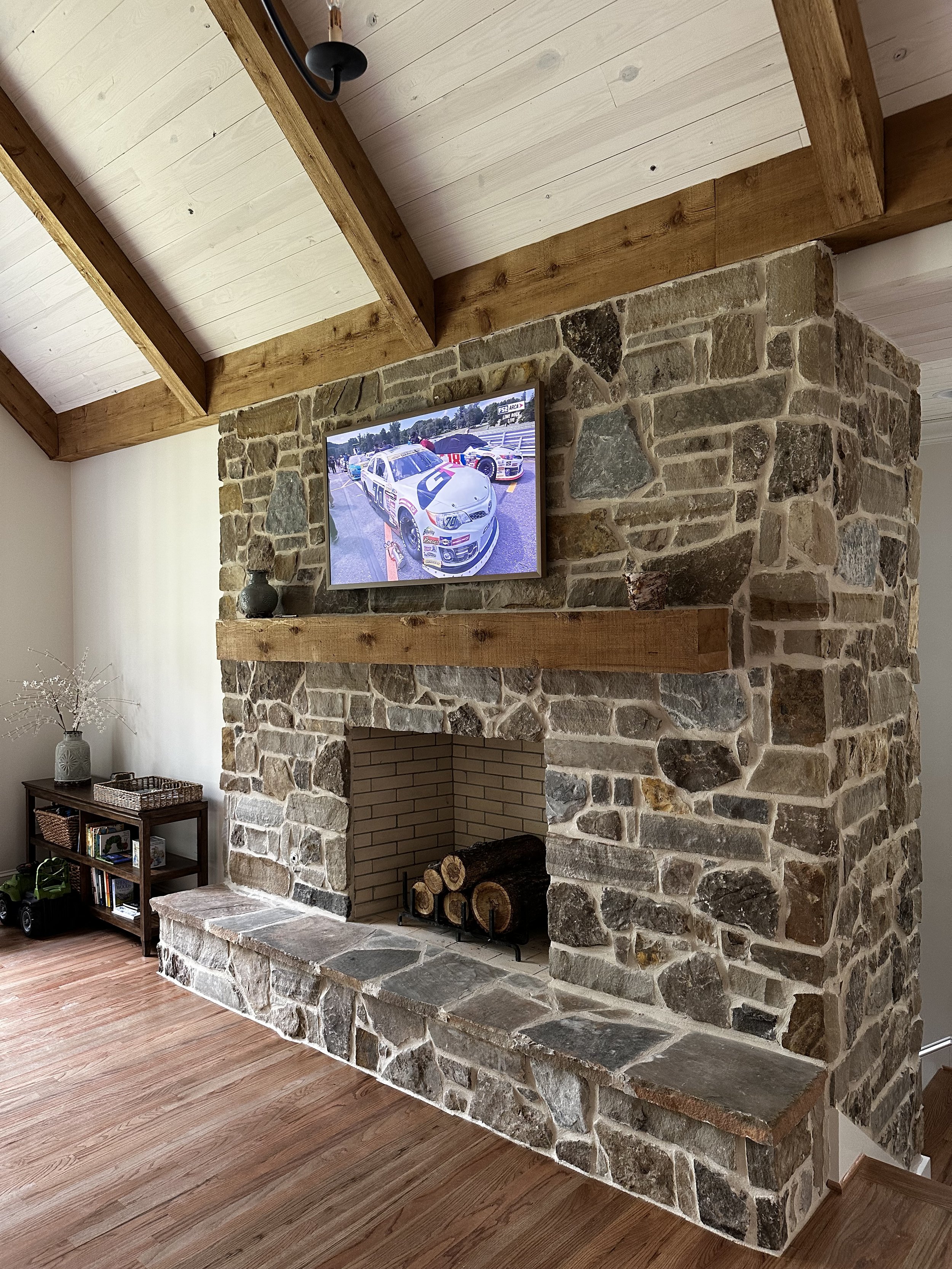Living room with a stone fireplace, wood mantle, and a TV mounted above it showing race cars. To the left, there is a small wooden bookshelf with decor and toys, and a vase with decorative branches.