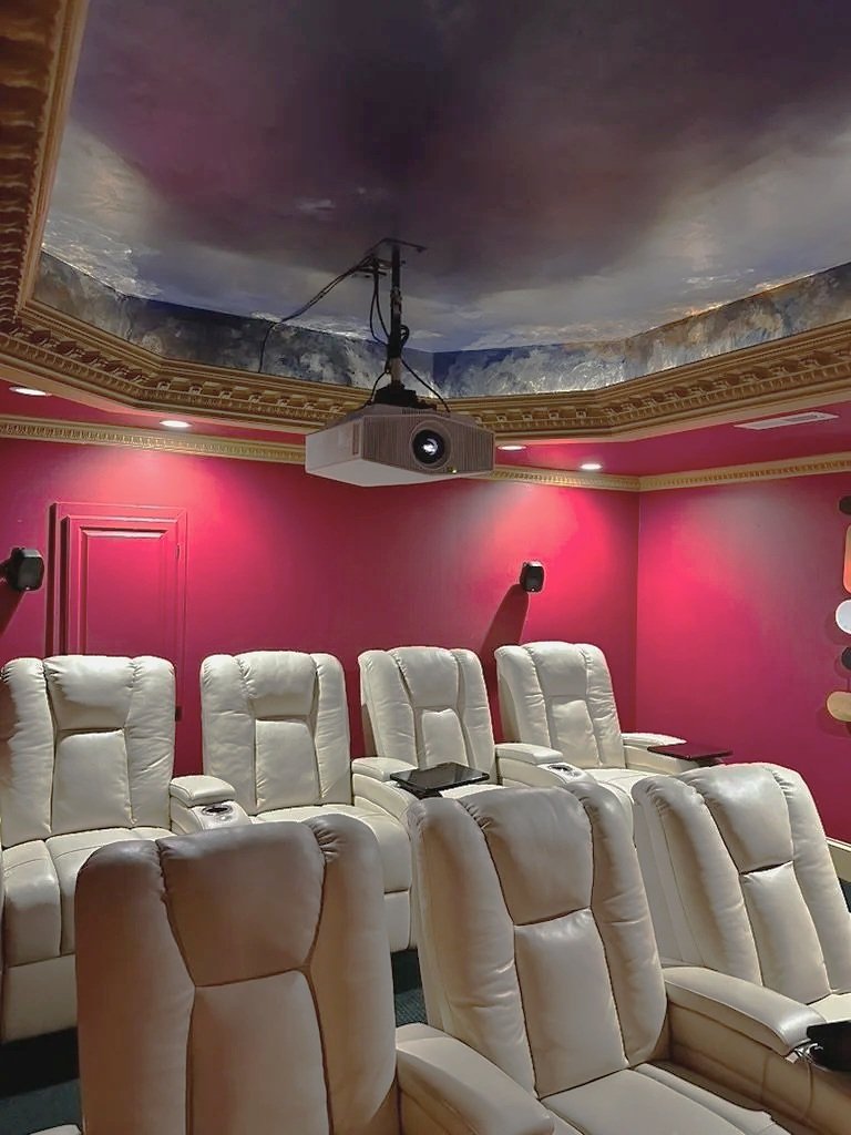 A private home theater with eight beige leather reclining chairs, a red wall with gold trim, and a ceiling projector mounted in the center.