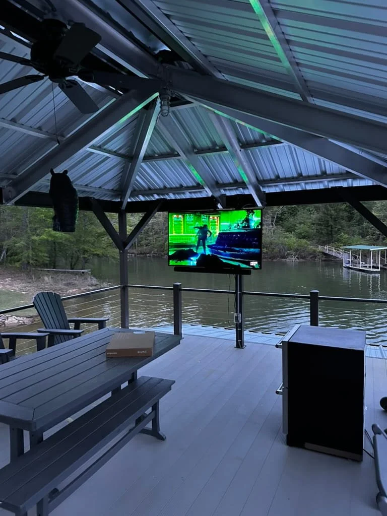 Outdoor pavilion by a river with a large TV, a ceiling fan, and seating area, illuminated with green LED lights.