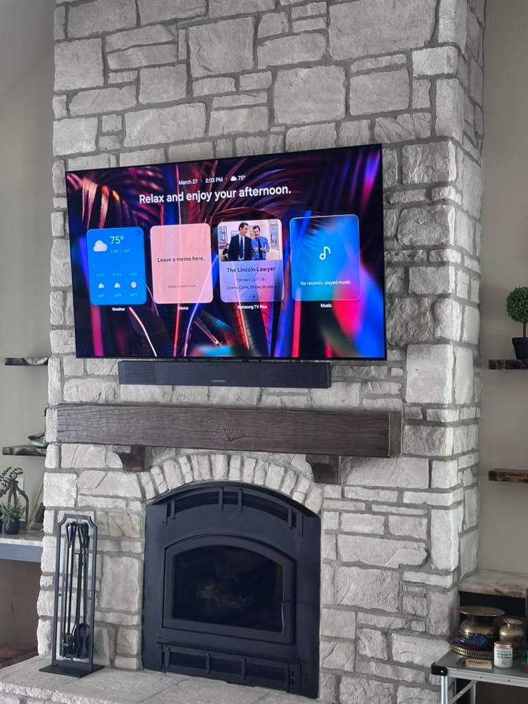 A flat-screen TV mounted above a stone fireplace with a wooden mantel, displaying a home screen with weather, notes, news, and music options in a living room.