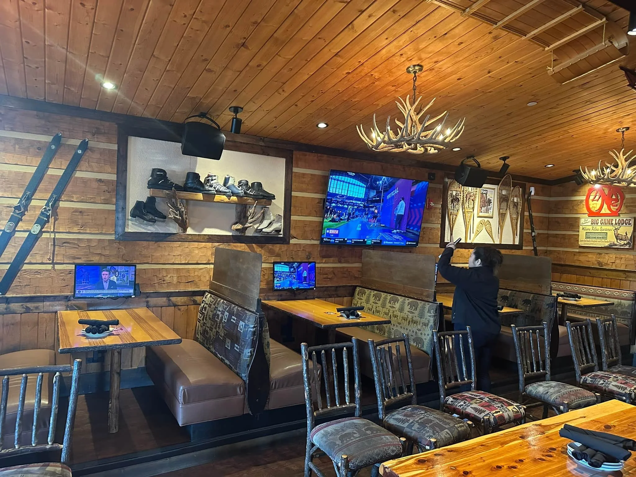 A restaurant or lodge interior with wood-paneled ceiling and walls, featuring antler chandeliers, multiple small TVs, shelving with shoes and snowshoes, and several tables and chairs, with a person in dark clothing standing and pointing at something.