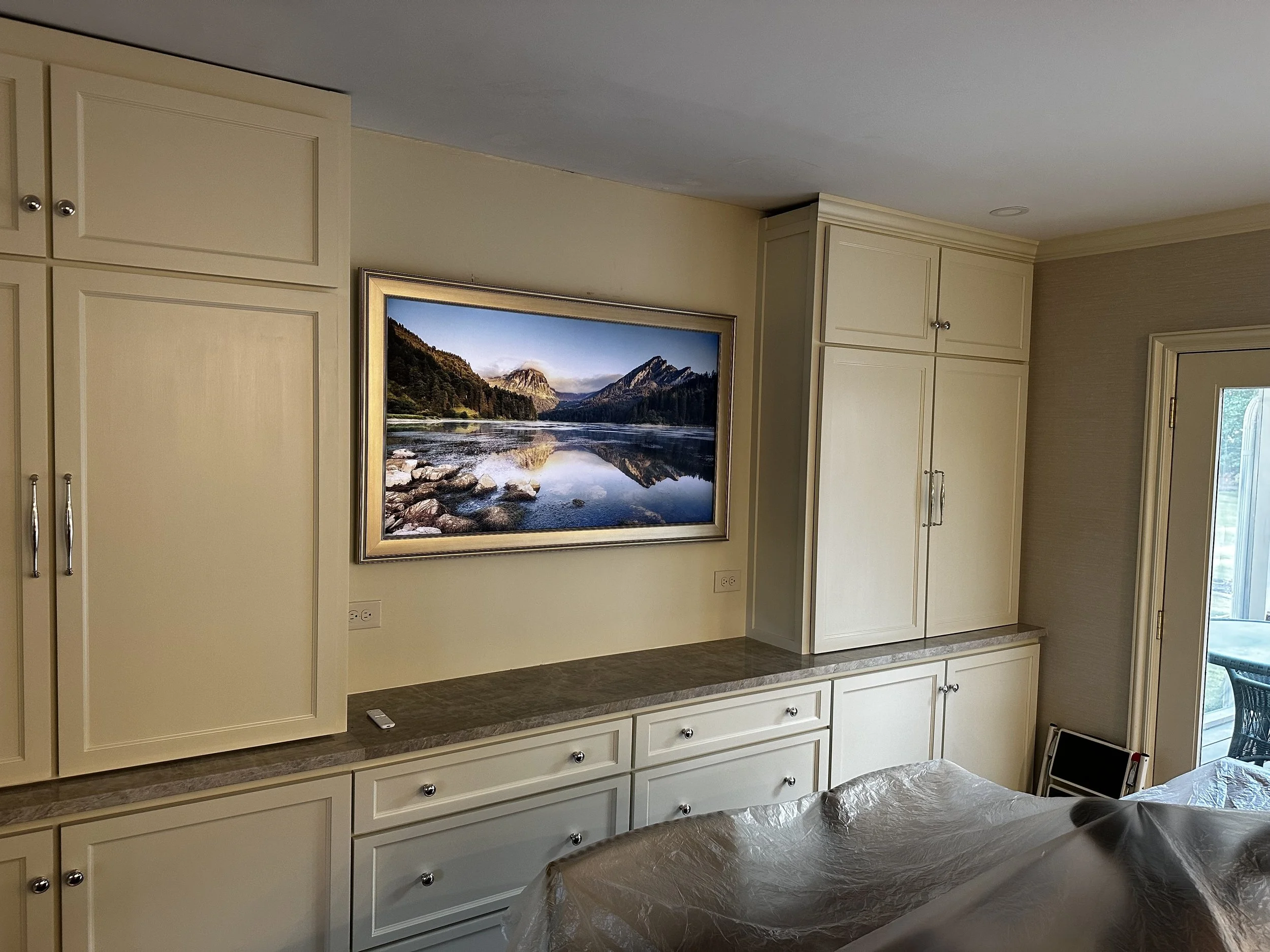 A framed landscape painting of mountains, trees, and a lake hangs on a beige wall above a built-in cabinet in a living room. The cabinet has cream-colored doors and drawers with silver knobs. To the right, there is a glass door leading to an outdoor patio with a chair visible outside.