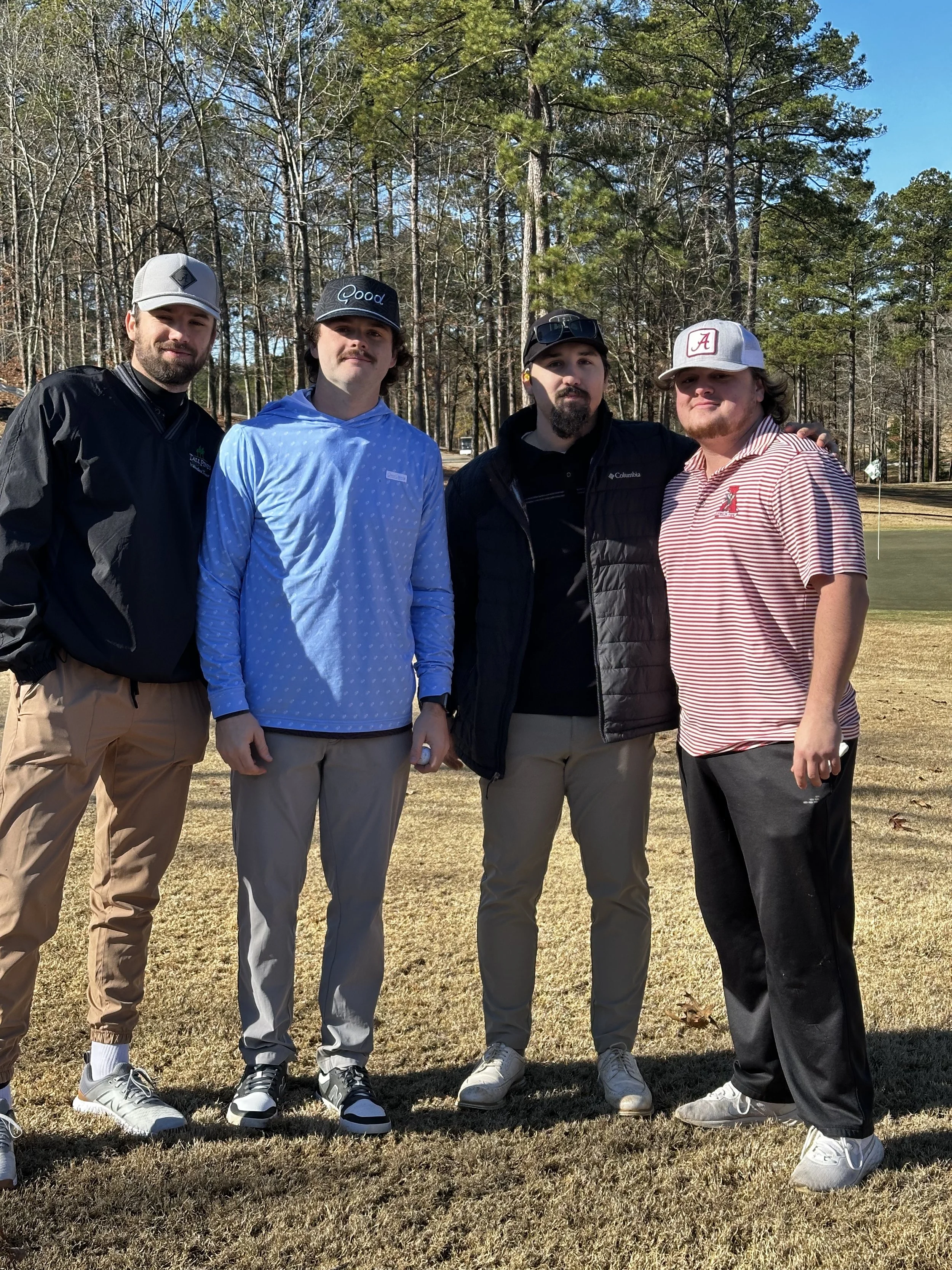 Four men standing on a grassy area with trees in the background, wearing casual attire and baseball caps, posing for a photo.