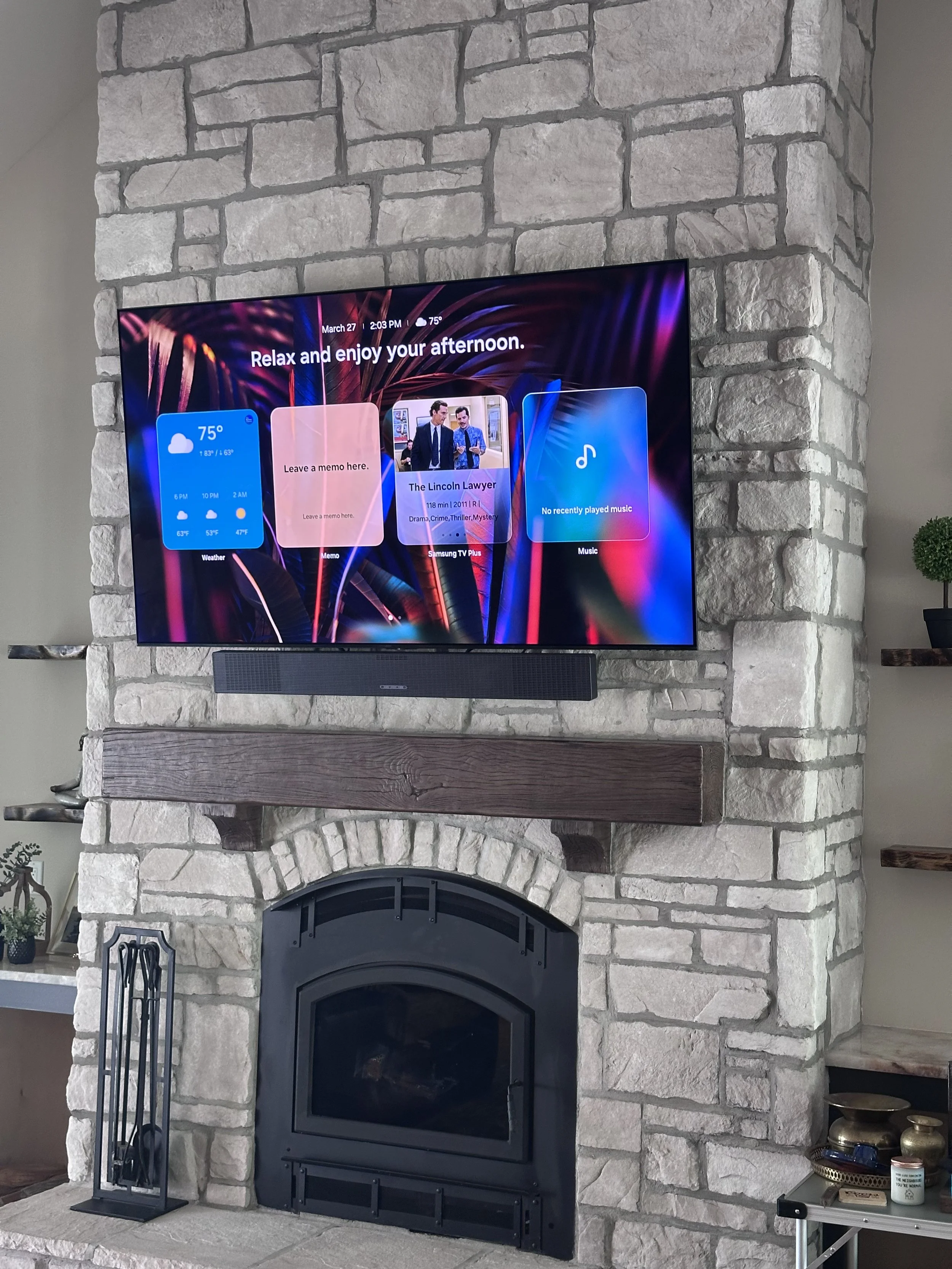 A stone fireplace with a wooden mantel and a flat-screen TV mounted above it. The TV screen displays a weather widget, a memo reminder, a TV show description, and music information. To the left of the fireplace, there is a fireplace screen with a decorative pattern. To the right, there is a small table holding decorative bowls and a jar, with a small potted plant visible in the background.