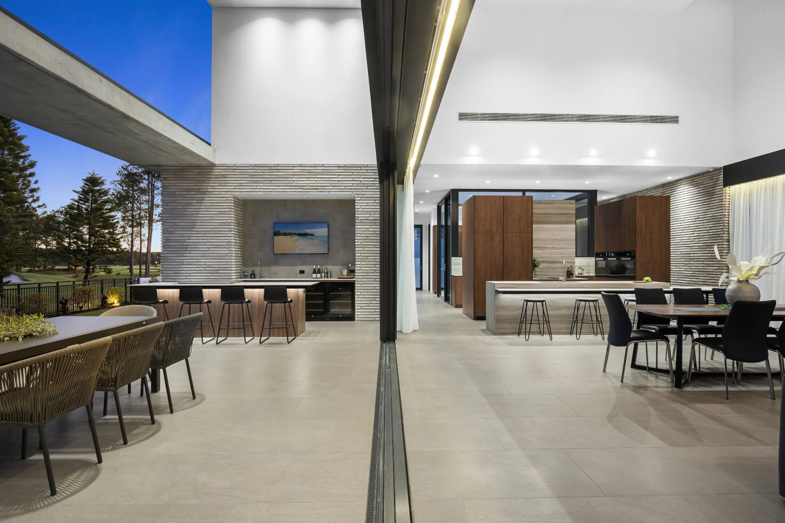 Modern luxury indoor-outdoor dining and entertainment space with open kitchen and bar area, featuring minimalist decor, sleek furniture, and large windows overlooking an outdoor landscape.